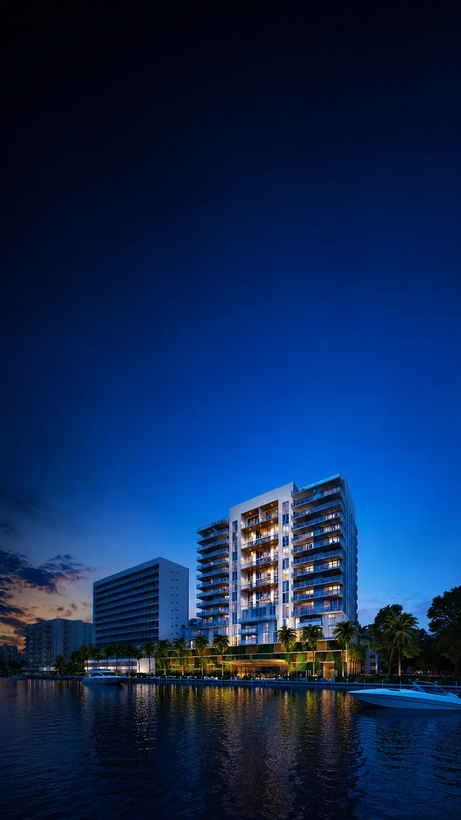 Waterfront residential tower in North Bay Village lit up against a deep blue evening sky, reflected in Biscayne Bay with docked boats and palm trees along the shoreline.