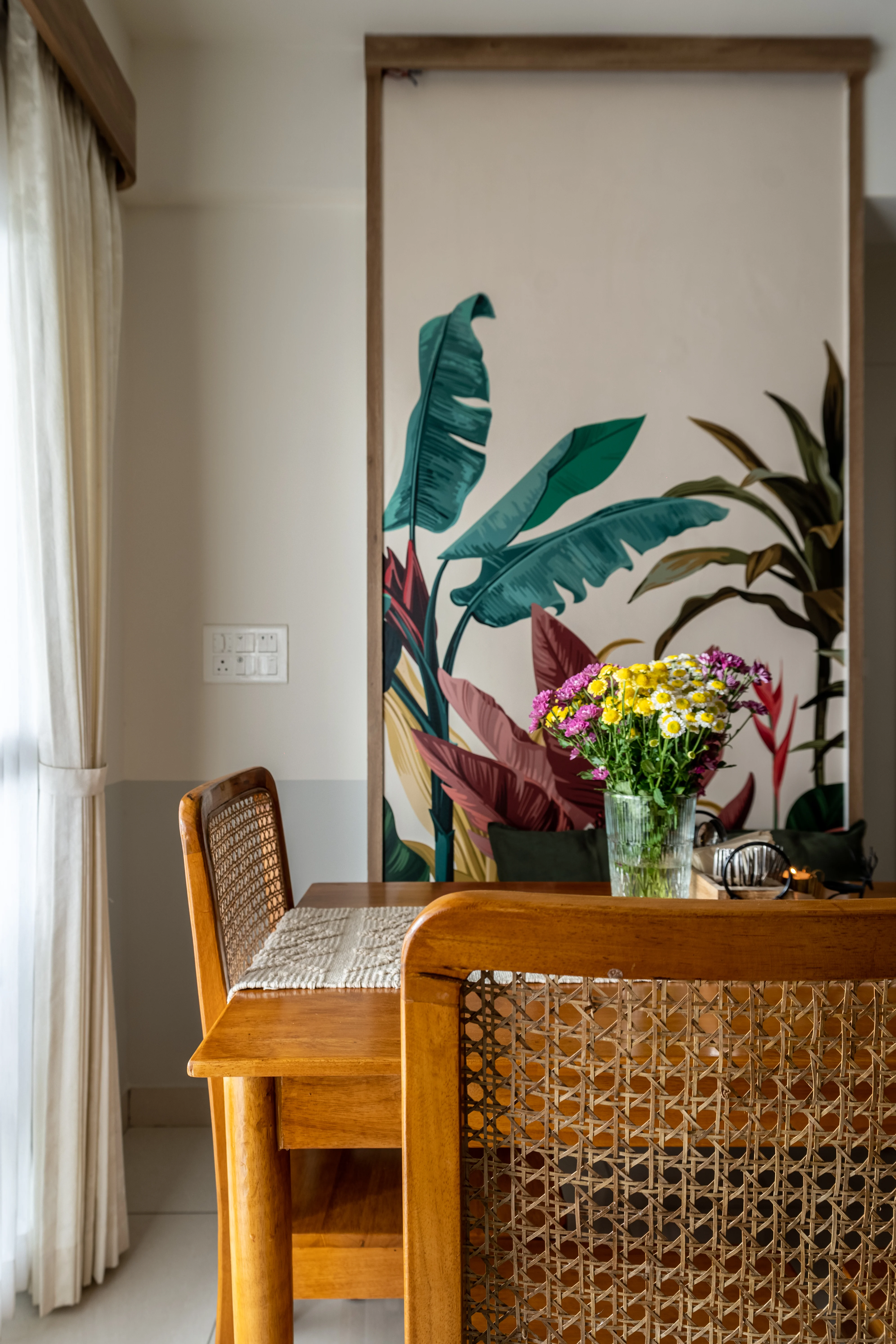 A close-up of a dining table with wooden chairs featuring cane woven backrests. A vase of colorful flowers sits on the table, and a large tropical-themed wallpaper covers the wall behind.