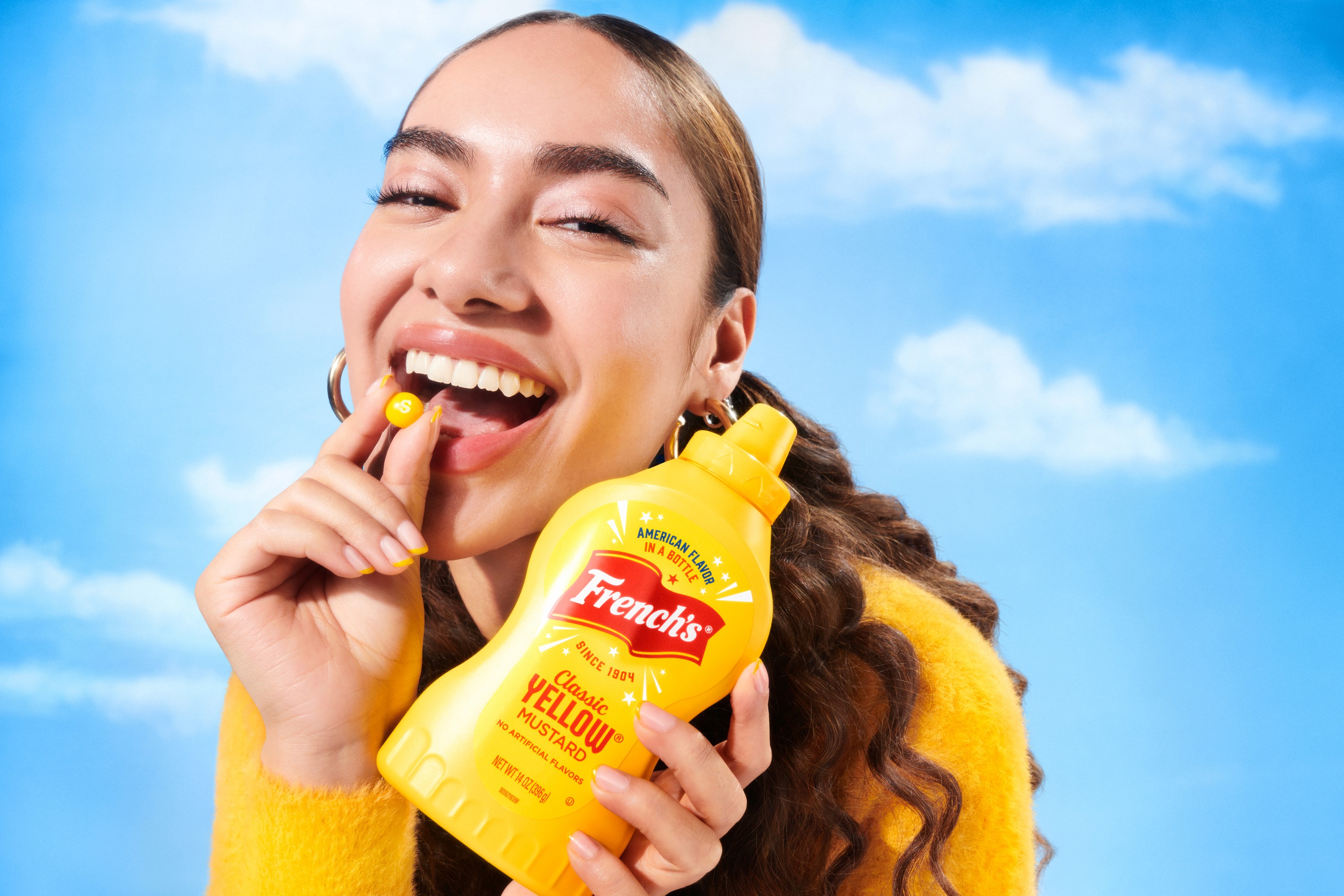 A smiling person in a yellow top holds a bottle of French’s Mustard against a bright sky with clouds.