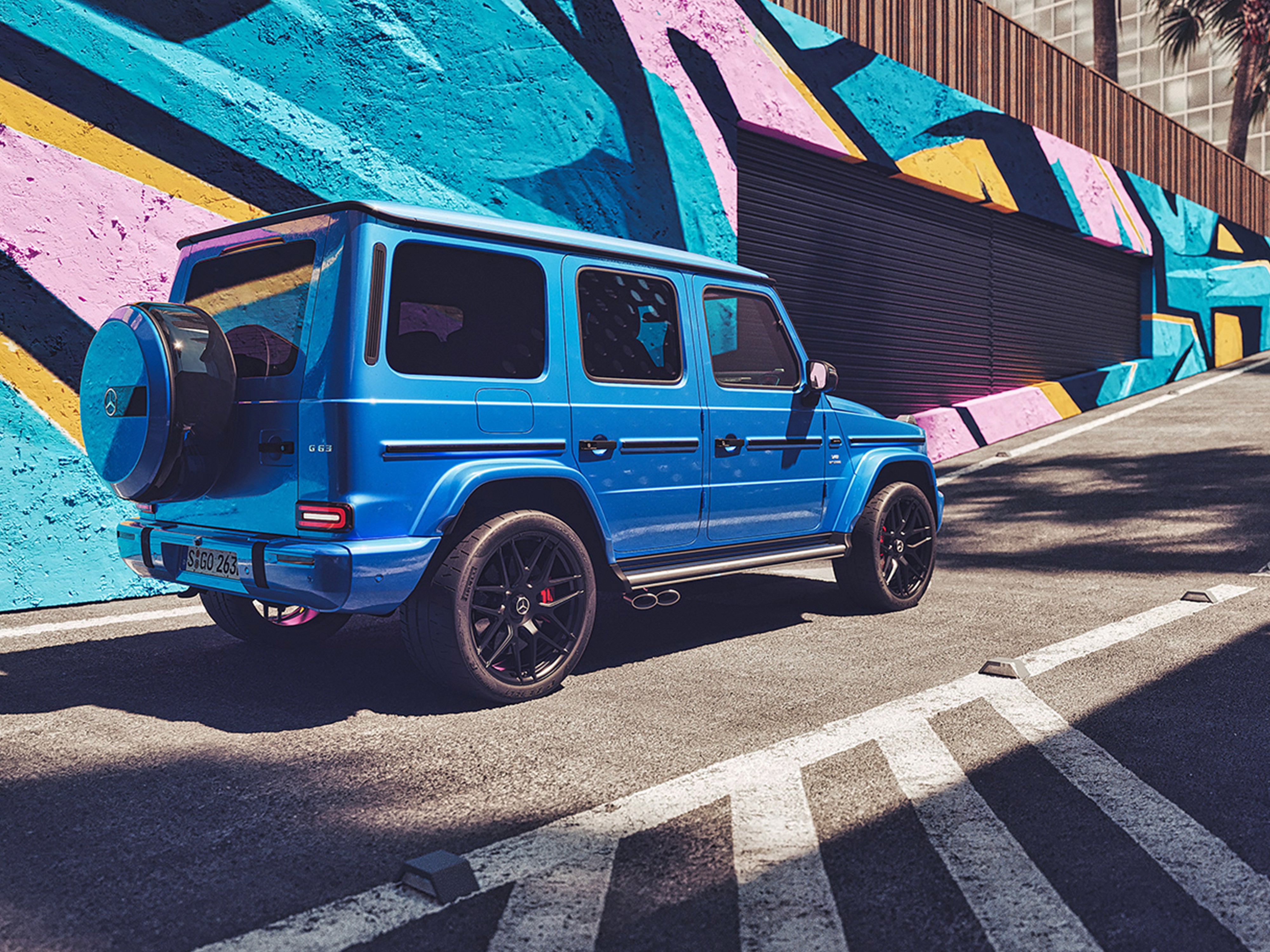 Render della Mercedes-AMG G 63 SUV di lusso blu brillante vista tre quarti posteriore parcheggiato davanti a un muro con graffiti colorati.