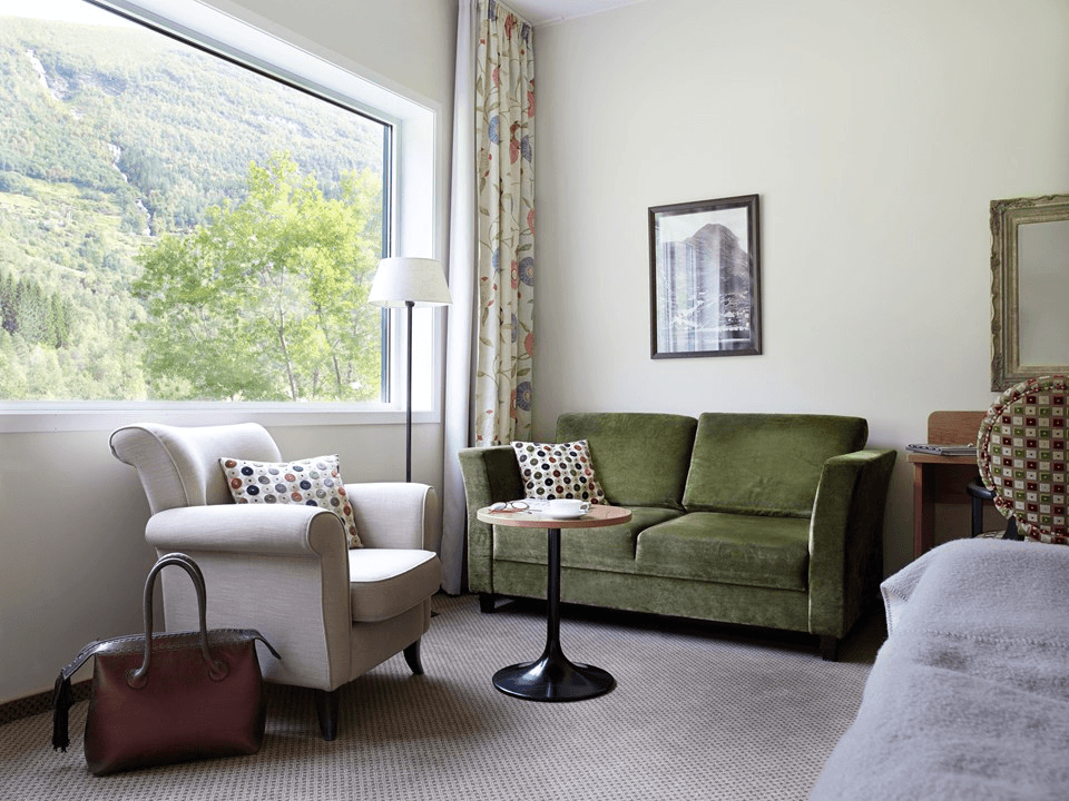A cozy living room features a plush green sofa and an inviting armchair beside a large window that provides a scenic view of lush greenery and distant hills.