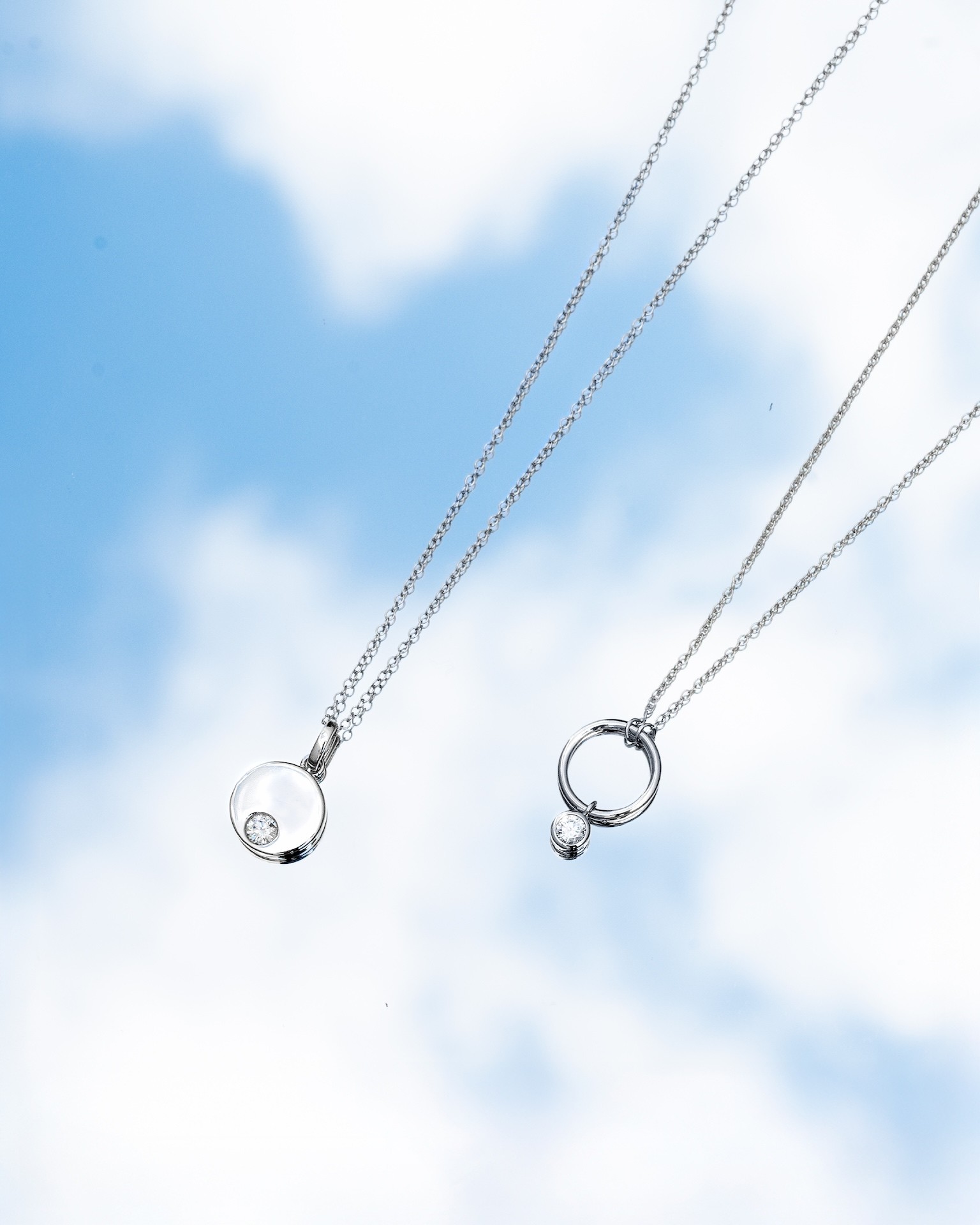 two silver necklaces with pendants sit on a relfective mirror surface, showcasing the blue sky and clouds as the reflected backdrop