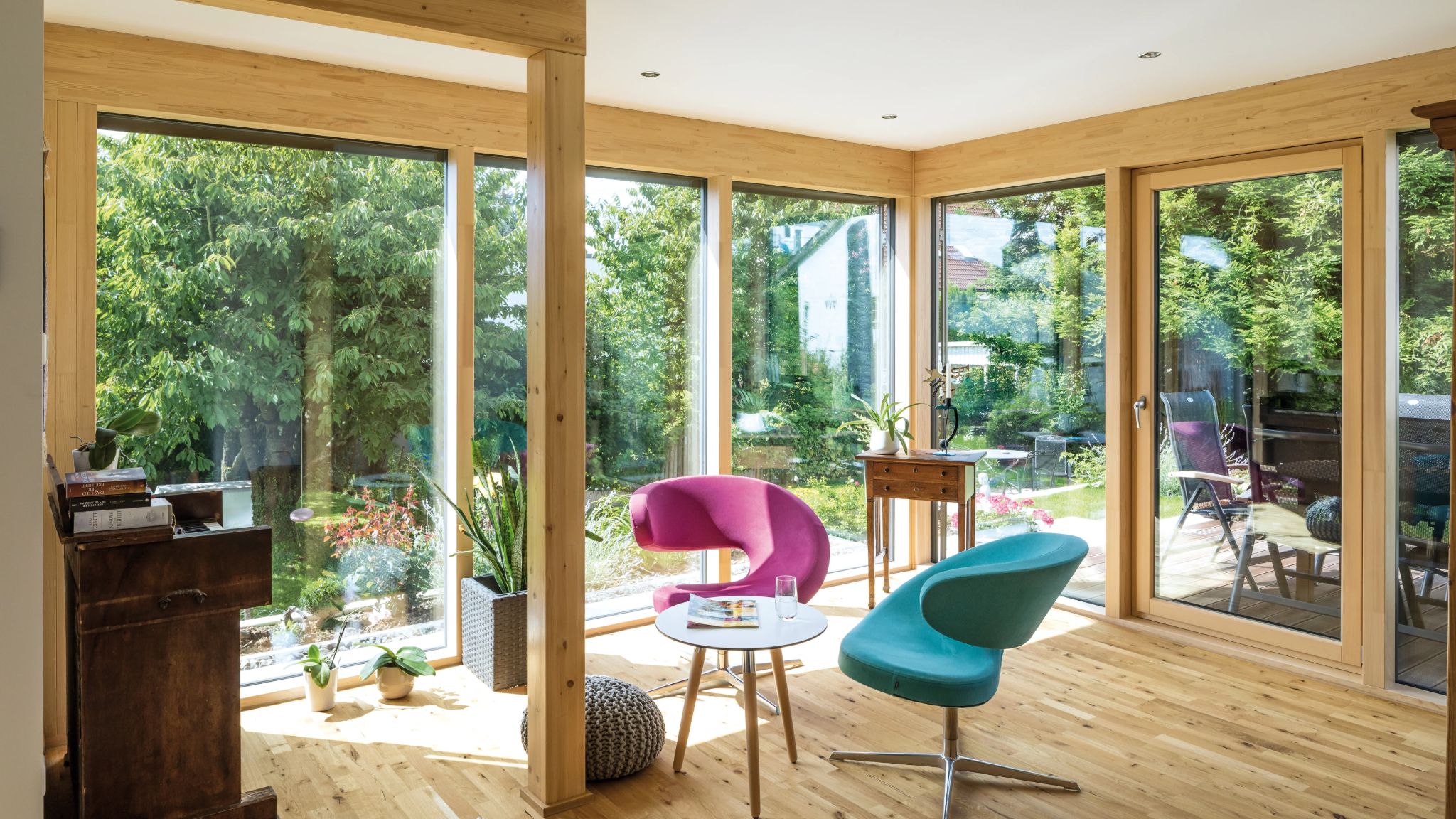Moderne Wohnzimmersitzgelegenheiten mit pinken und türkisen Stühlen, umgeben von großen Fenstern mit Blick auf einen üppigen Garten.