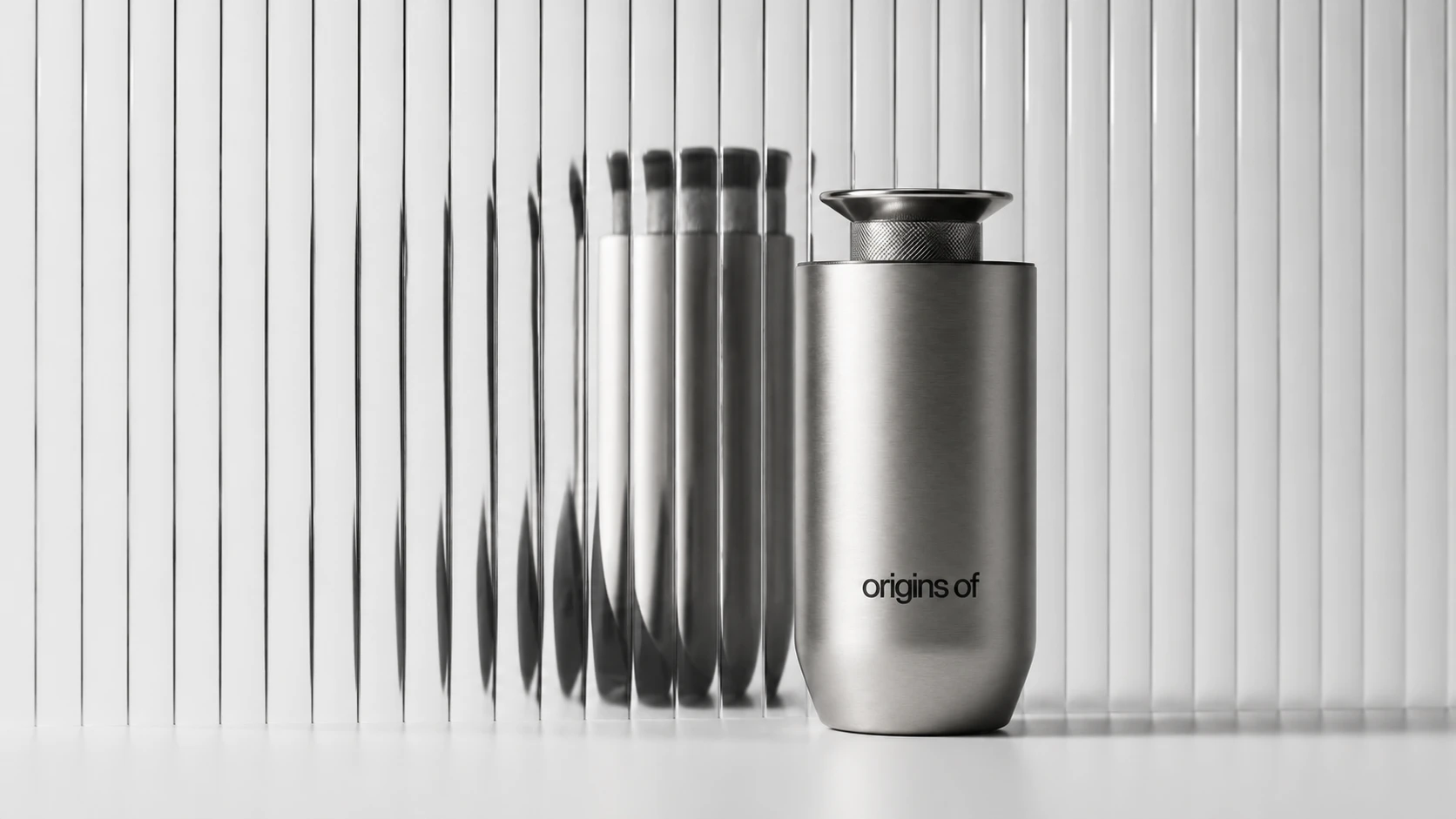 A brushed-steel Origins Of waterless diffuser with a knurled top standing on a white surface, its reflection repeated and distorted behind a pane of fluted glass.