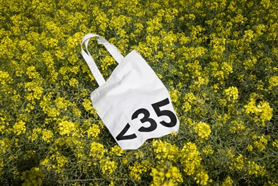 White tote bag with Less Than 35 bold typography among yellow flowers — cultural branding by OUF Design Studio