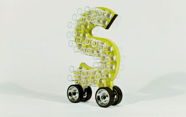A light yellow, three-dimensional letter “S” is constructed from transparent plastic bottles arranged in rows. The “S” has four black wheels attached to its base. The background is plain white.