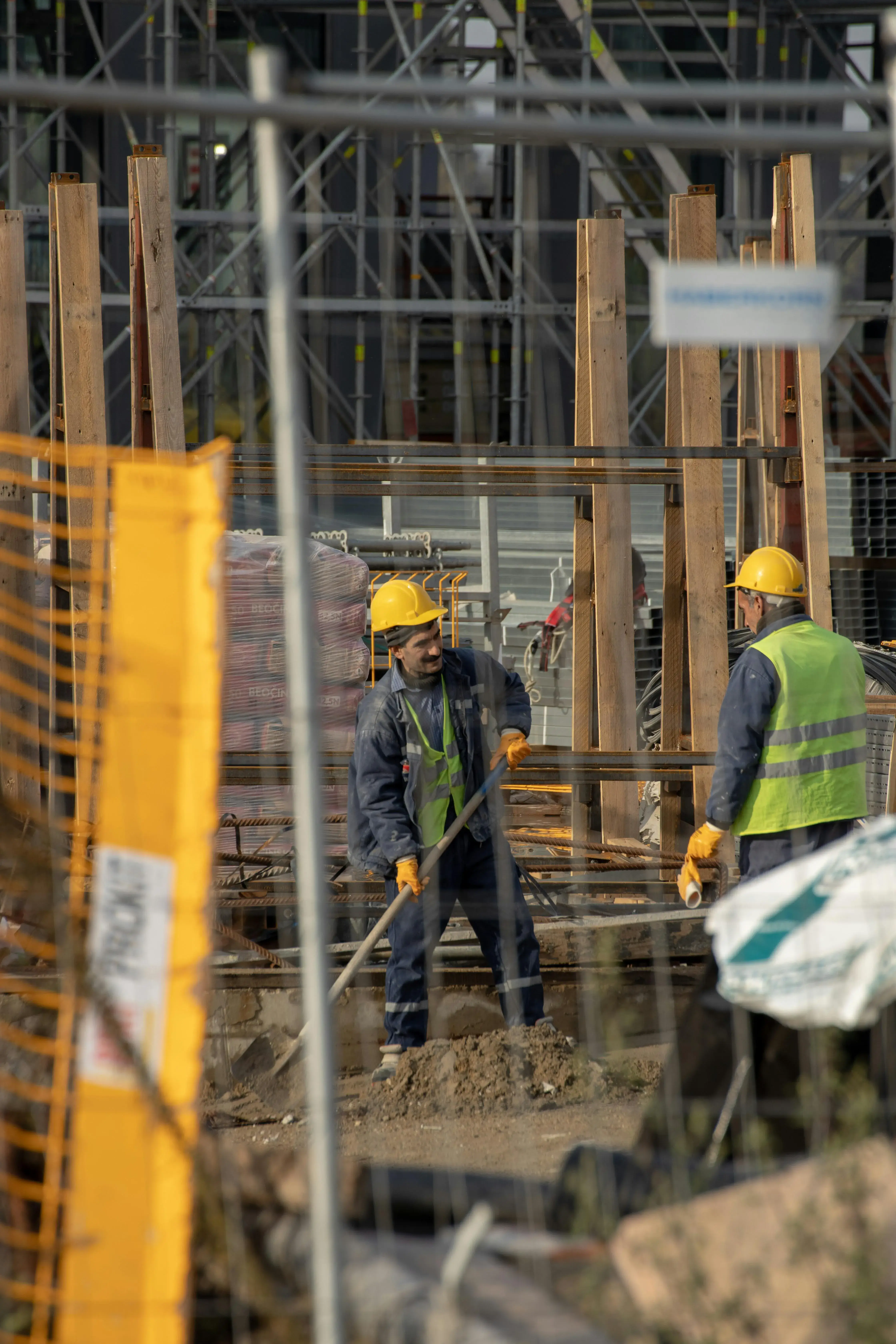 Two construction workers in hard hats and vests stand on a site with scaffolding, surrounded by wood beams and fencing, engaging in conversation.