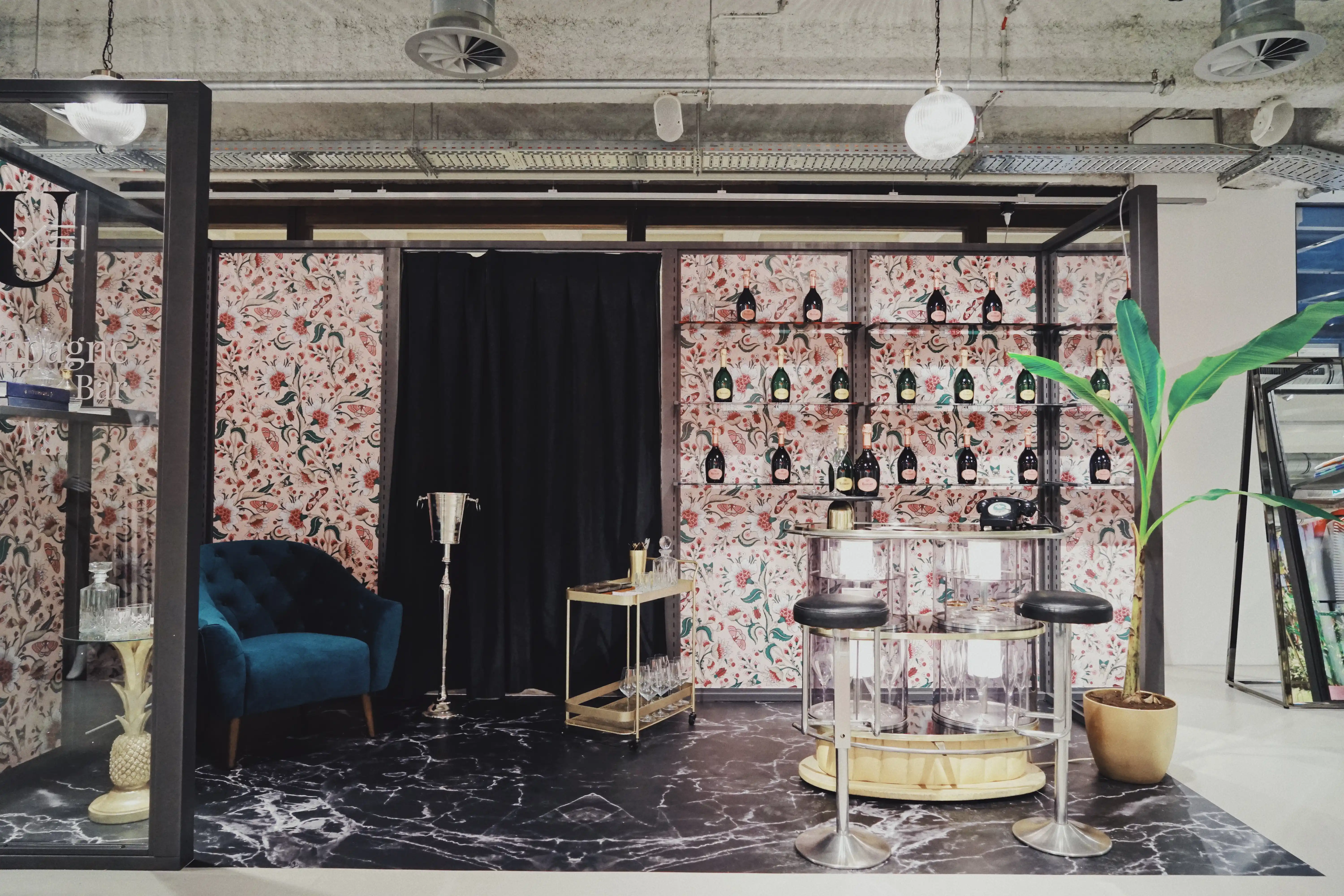Champagne lounge at UZWEI FASHION HOTEL with floral wallpaper, bottle shelves, a teal velvet sofa, a silver ice bucket on a stand, a gold bar cart and a clear acrylic bar with stools.