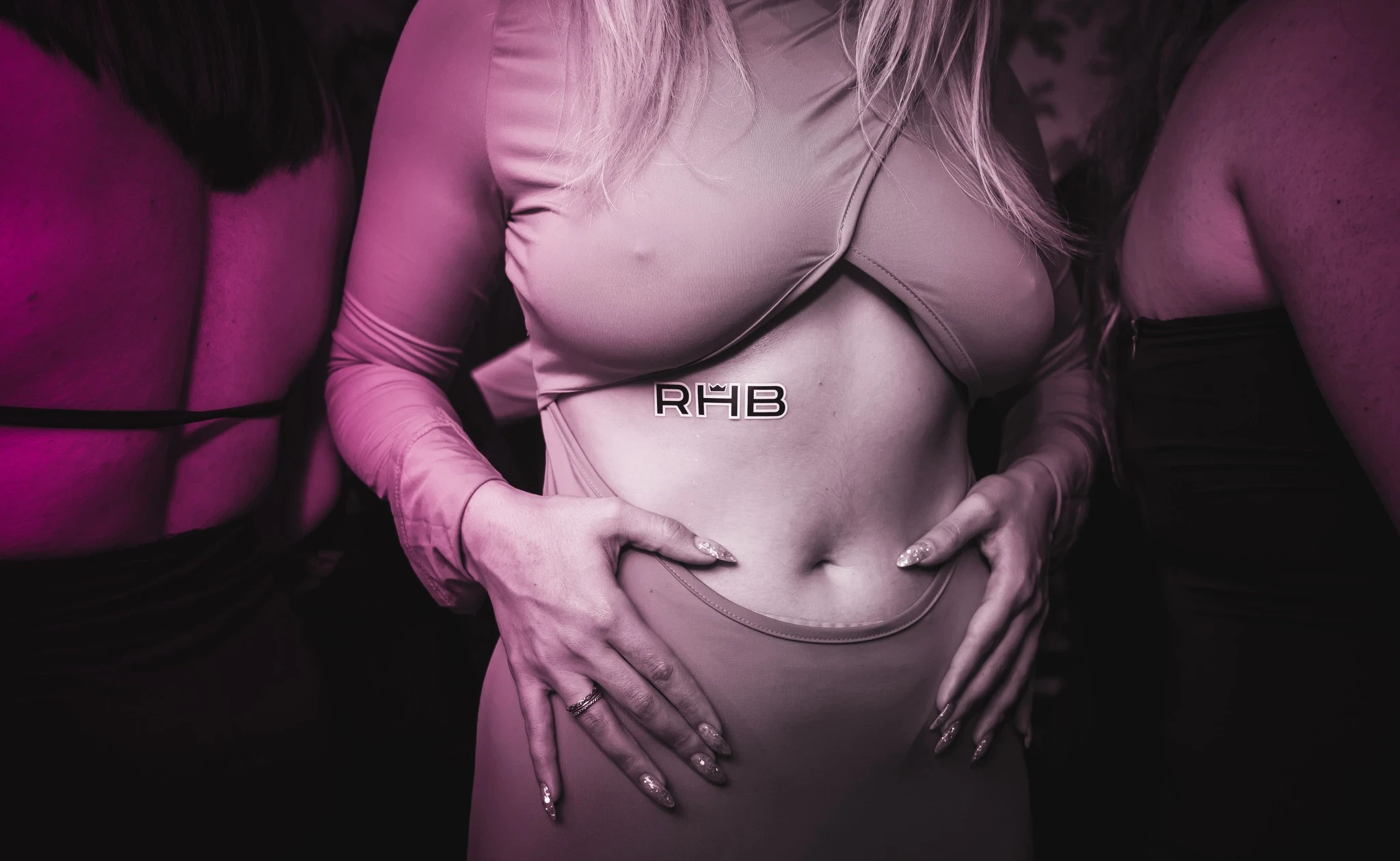 A close-up of a person's midsection at the RHB party, wearing a light-colored cutout dress. Their hands, with long glittery nails, are on their hips, and an "RHB" sticker is on their stomach.