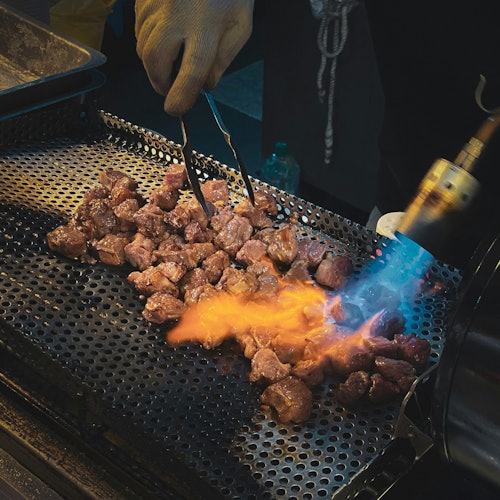 一个人用钳子和可见火焰的喷灯在有孔的烤架上烤肉块。