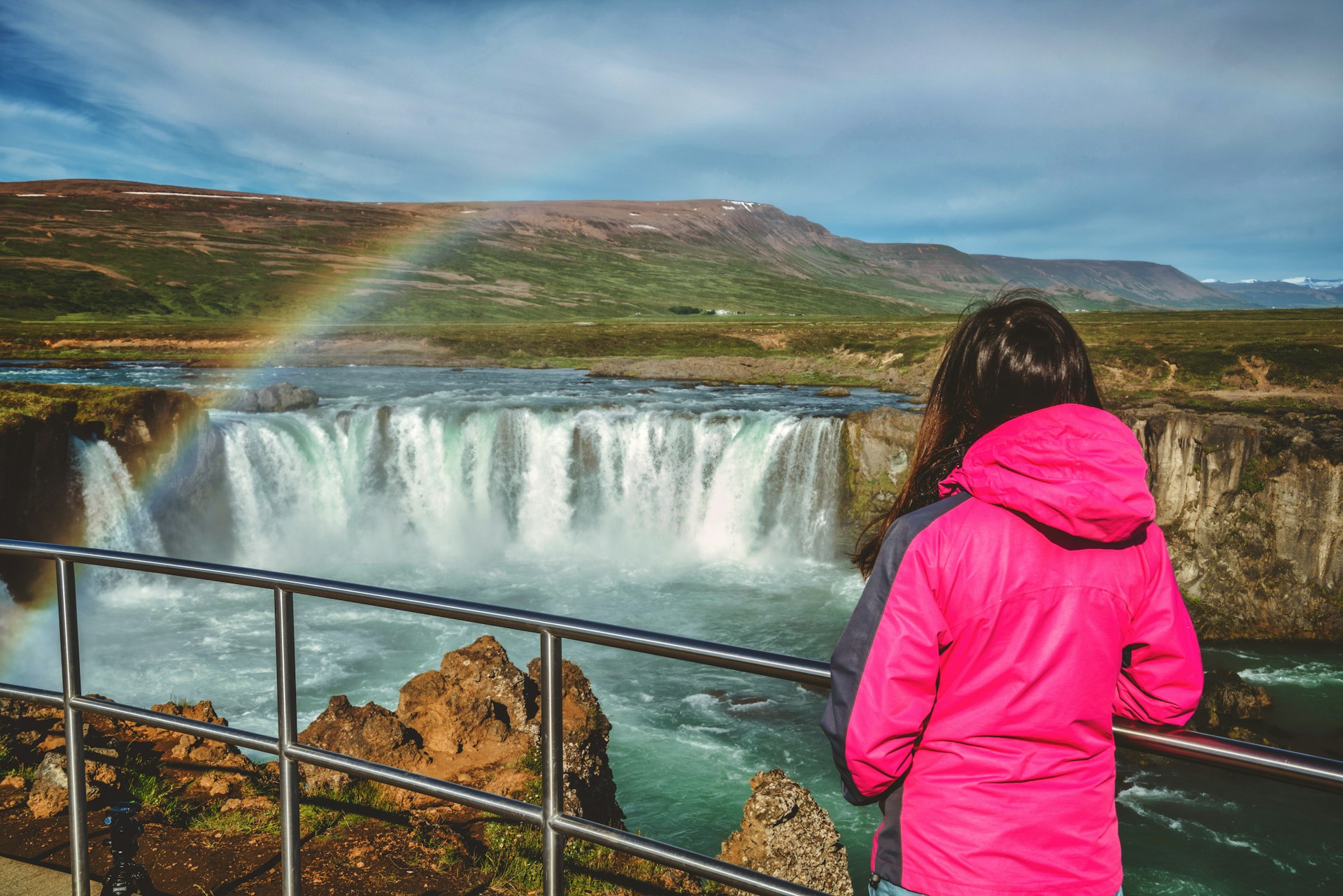 Visitor overlooking Godafoss Waterfall with a rainbow in North Iceland.
