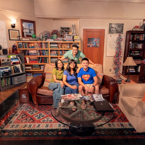 Four people sit on a couch in a living room filled with bookshelves, scientific models, and various decor; a coffee table is in front.