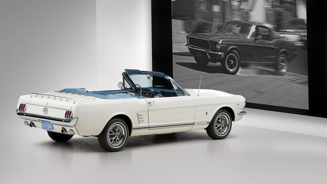 A white Ford Mustang convertible with a blue interior is positioned on a light gray floor, facing left. A large black and white photograph of a black Ford Mustang is mounted on a black frame to the right of the car. The license plate on the white Mustang reads “119 048”. The background is plain white.