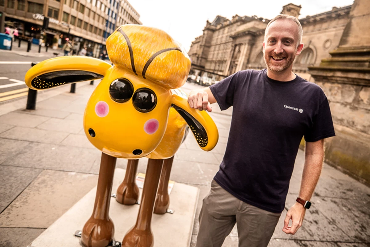 A person stands next to a large yellow Shaun the Sheep sculpture on a city street, lightly touching its ear.