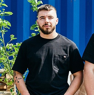 A person stands in front of a blue wall, wearing a black t-shirt, with green plants in the background.
