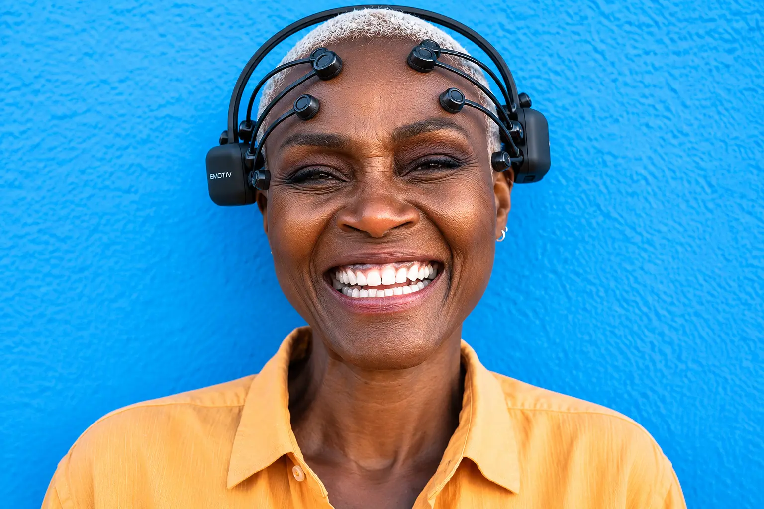 A smiling African woman with short white hair wears an Emotiv Epoc X headset