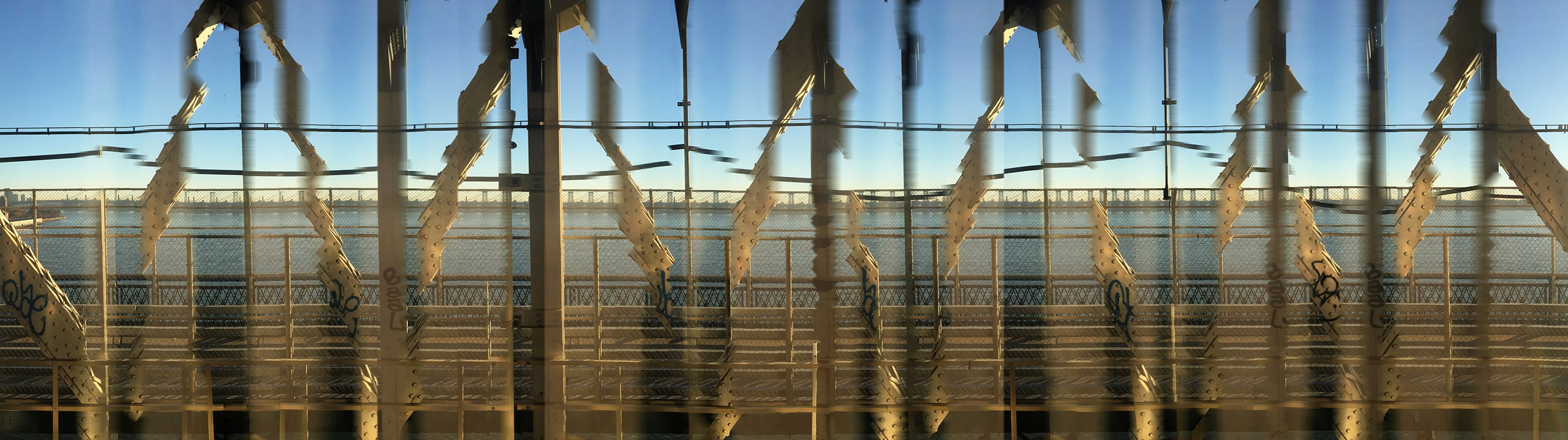 Panoramic photograph of a bridge crossing over water at sunset, with repeating steel beams and railings stretched into vertical distortions. Warm light and rhythmic patterns create a dreamlike sense of movement through space and time.