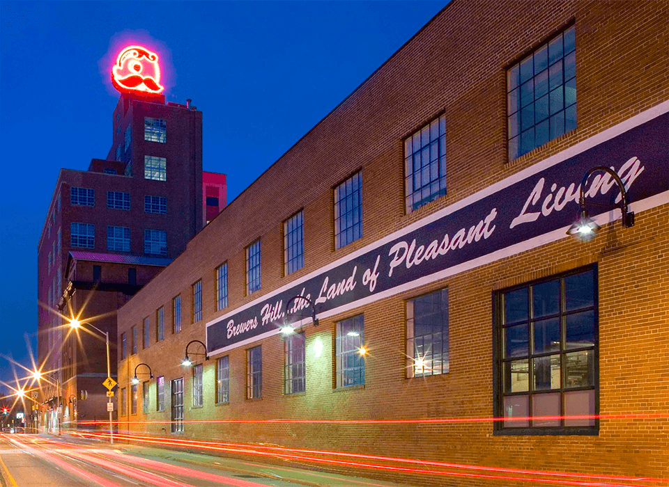 Two women wearing aprons stand in a kitchen with exposed brick walls, holding dough and smiling at the camera—looking like media pros collaborating in an agency environment. Cooking utensils and pots hang on the wall behind them.