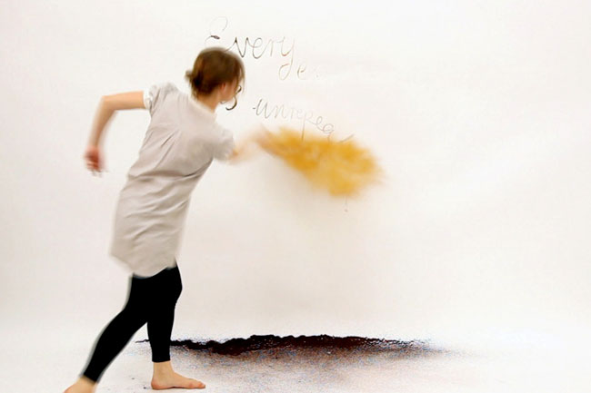 A person with light brown hair, wearing a light gray short-sleeved shirt and black pants, is captured mid-motion against a white wall. Their right arm is extended forward, and their left arm is bent at the elbow. A yellow, cloud-like shape is visible near their right hand. The ground beneath them is dark and textured. The words “Everyday unrep” are written on the wall behind them.