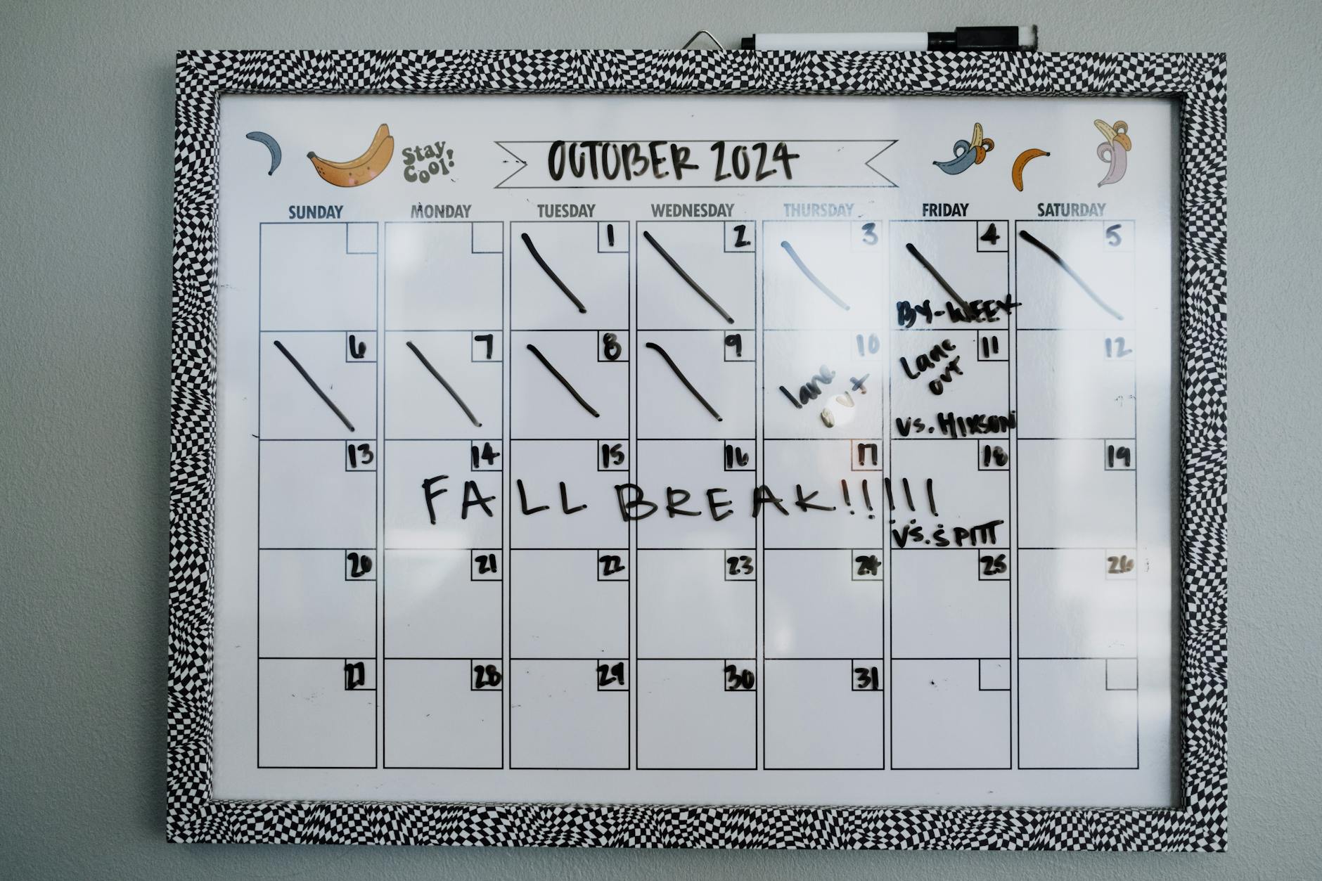 A hand checking off boxes on a printed weekly schedule pinned to a corkboard next to a classroom door.