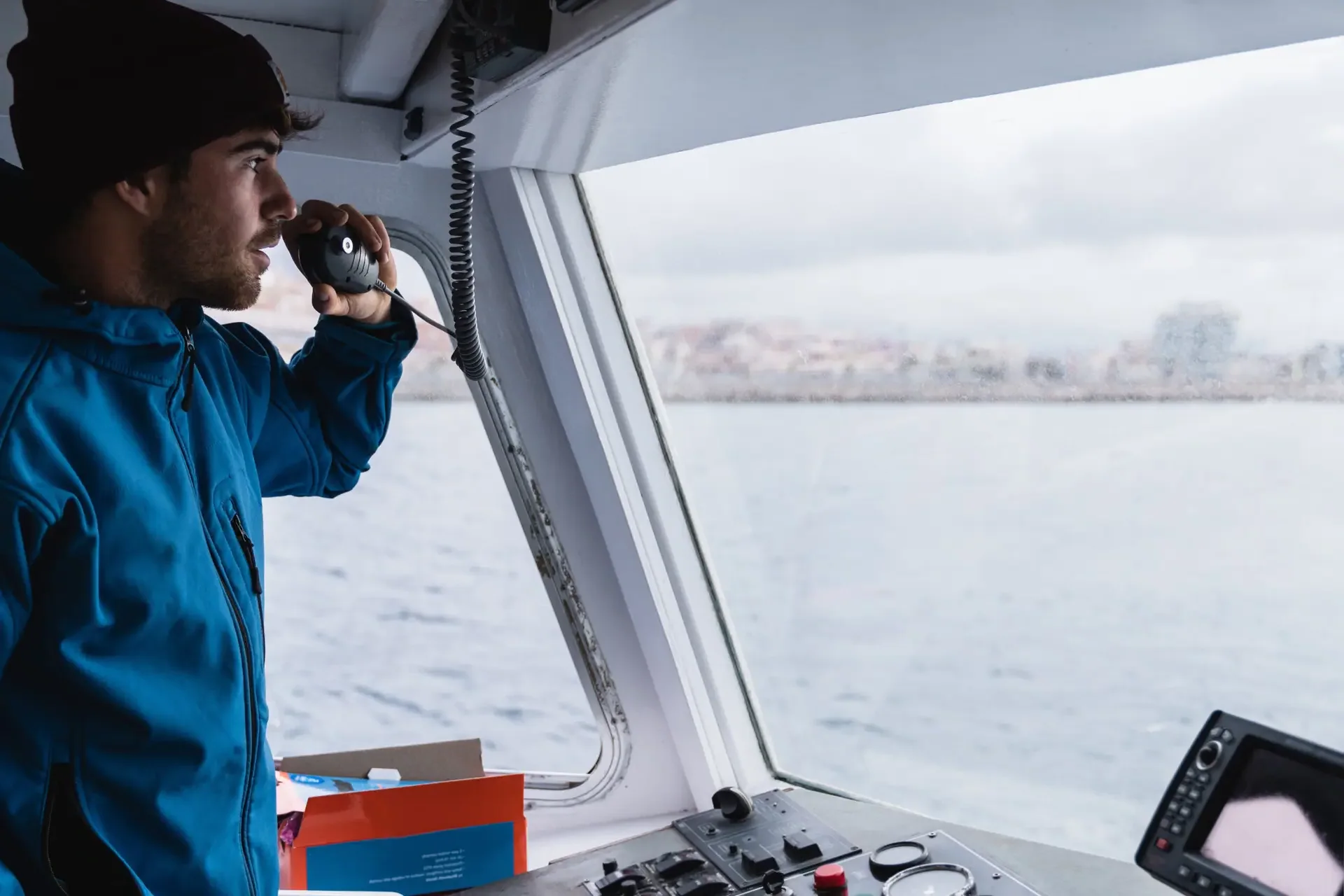 Skipper im Steuerhaus spricht in das Handmikrofon der Seefunkanlage, im Hintergrund die Küste