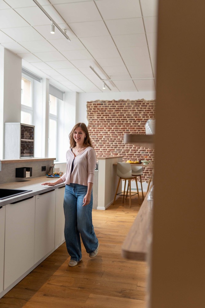 Mitarbeiterin steht in einer Büroküche und guckt in die Kamera, im Hintergrund Regale und Backsteinwand.