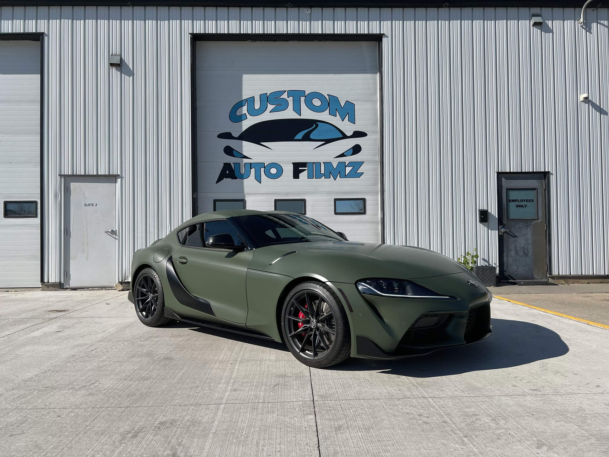 A matte green Toyota Supra sports car is parked on a concrete driveway in front of a white industrial building.