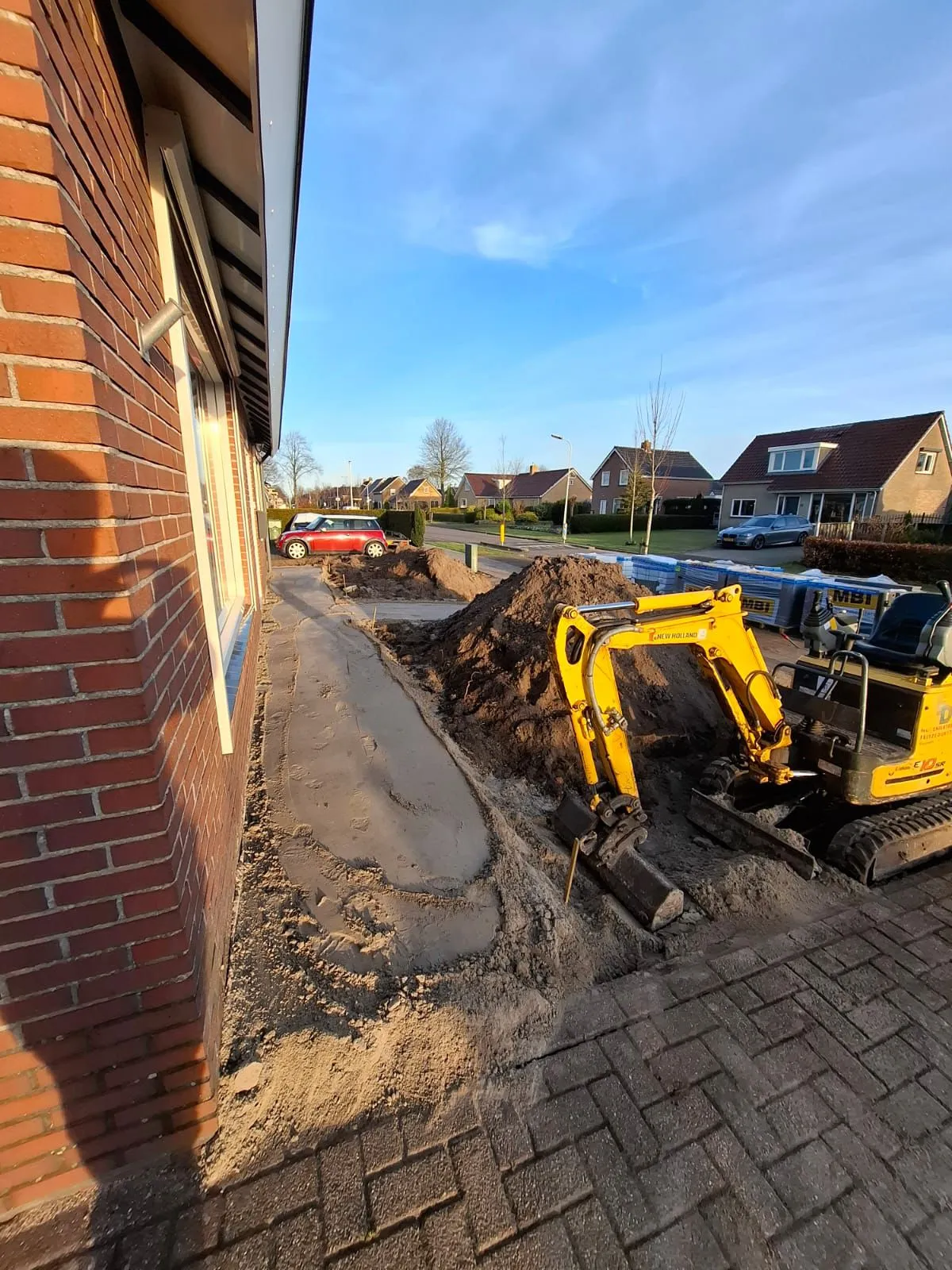 Mini graafmachine werkt aan het voorbereiden van een onderhoudsarme voortuin met nieuw pad naast een woning in Harkema.