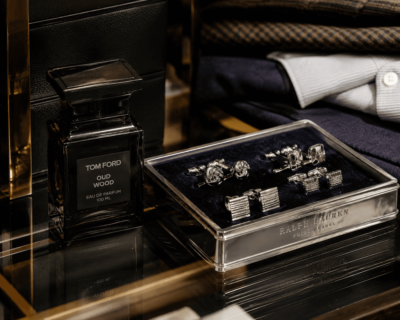 Close-up of luxury men's grooming accessories on a glass shelf, featuring a 100ml bottle of Tom Ford Oud Wood Eau de Parfum next to a silver Ralph Lauren Purple Label presentation box holding multiple pairs of classic silver cufflinks on a navy blue velvet lining, with folded shirts in the background.