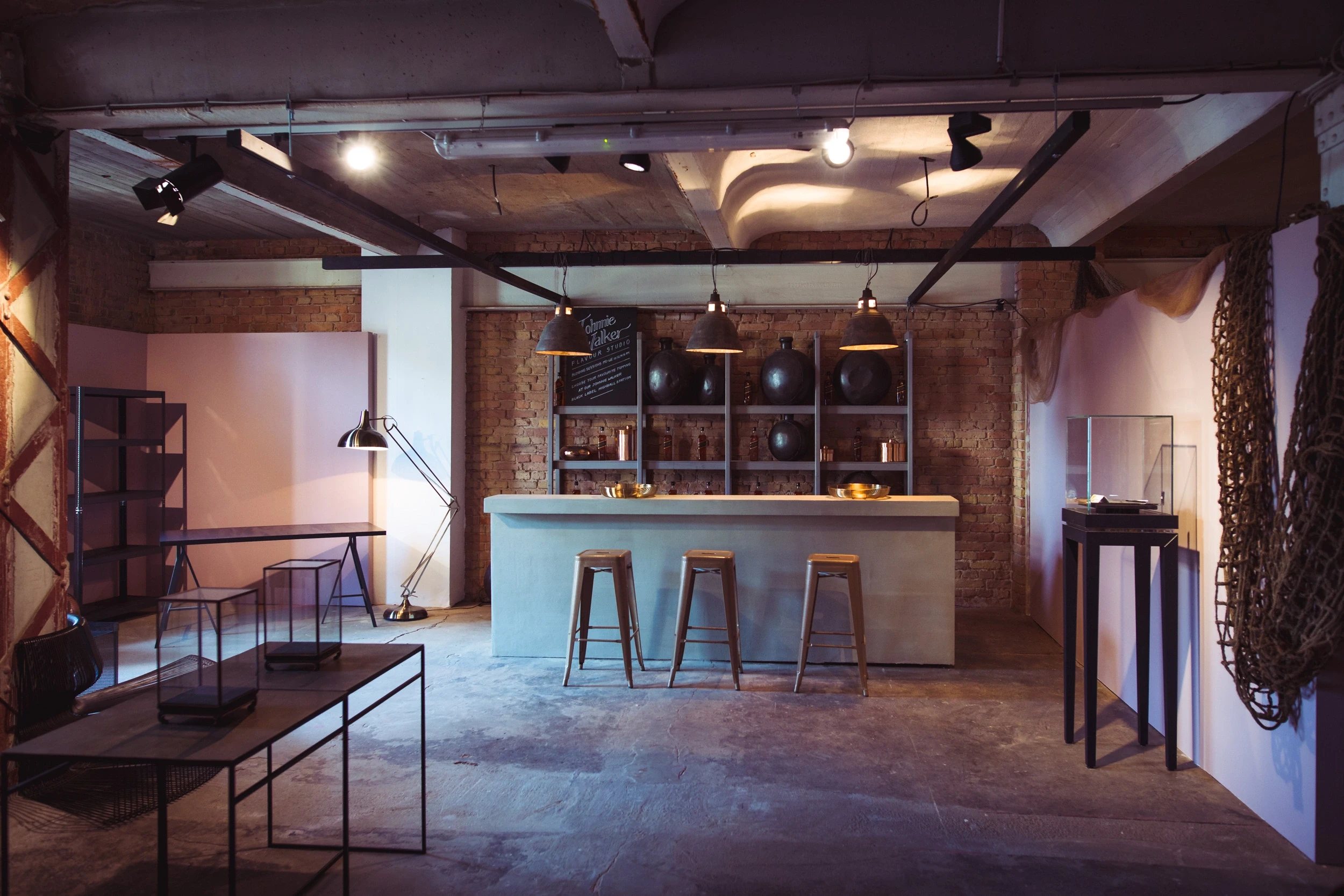 Concrete bar at DIAGEO BCB18 with wooden stools, shelving with black spherical vessels and copper pendant lamps against exposed brick.