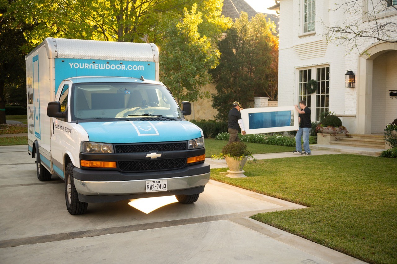 A blue and white delivery truck is parked on a concrete driveway in front of a suburban home, while two workers carry a large white and blue door towards the entrance, surrounded by lush green grass and shrubbery.