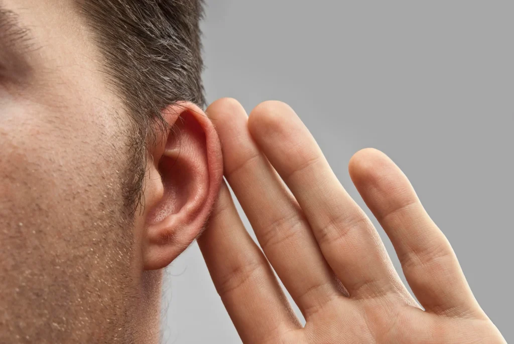Elderly patient wearing audiometric headphones during a hearing test, with an audiologist visible through the sound booth window