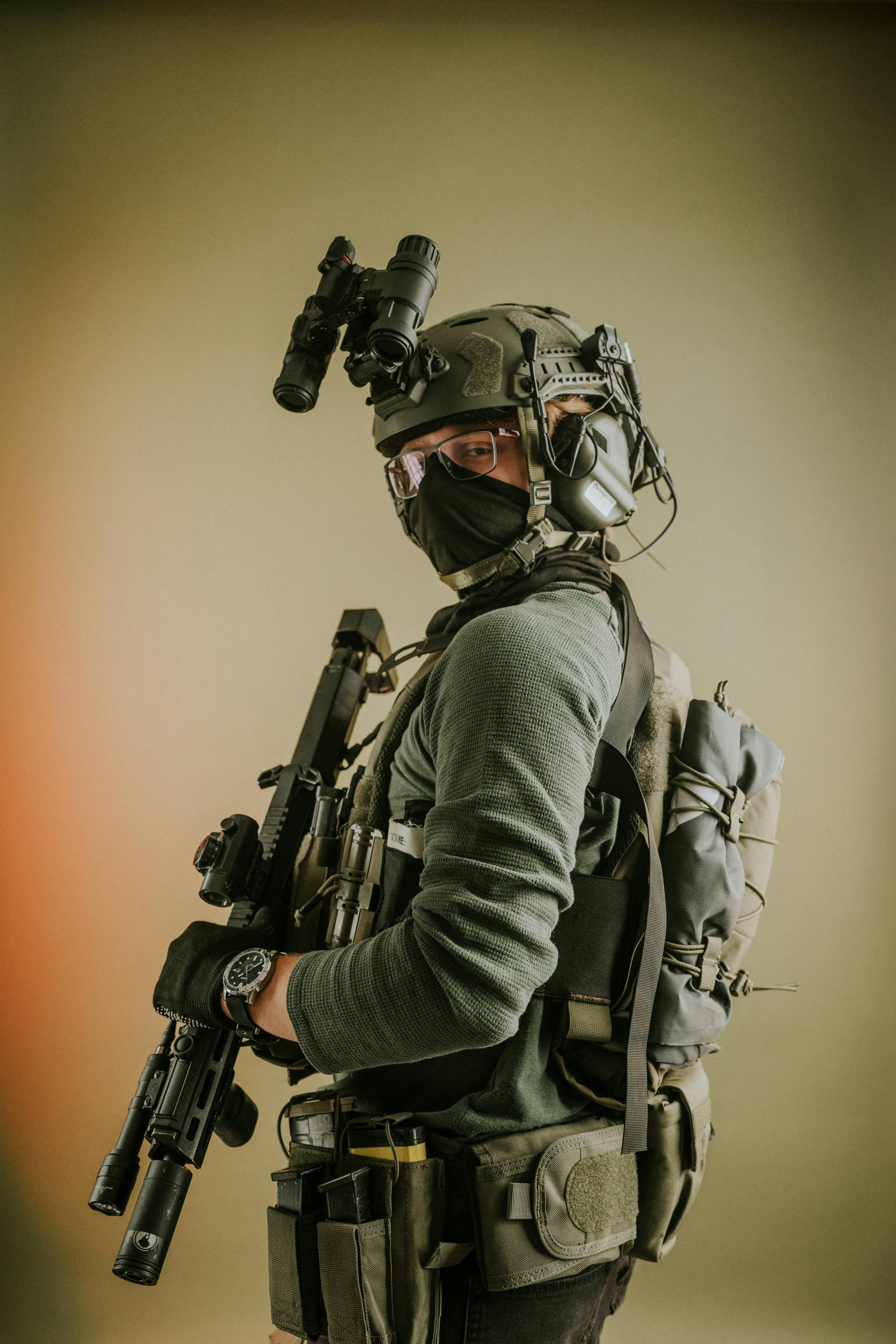 A tactical operator in full combat gear holding what appears to be a training weapon with a blue barrel tip (indicating it's likely a training or simulation firearm). The person is wearing a tactical helmet with face covering, body armor, and camouflage clothing in a kneeling position in what looks like an urban training environment with smoke or mist visible.