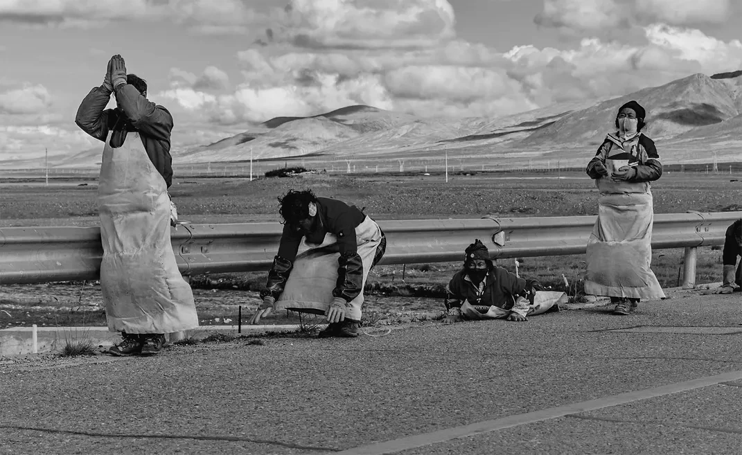 Black and white documentary photograph by Qinglin Li titled "Spiritual Pilgrimage." The image captures several Tibetan devotees performing full-body prostrations along a desolate highway on a high-altitude plateau. Each person is in a different stage of the ritual—one standing with hands in prayer above the head, others bowing or lying flat against the asphalt. They wear protective aprons and hand pads for the grueling journey. The vast, rugged mountains of Tibet and a cloud-filled sky stretch across the background, emphasizing a sense of profound devotion and spiritual resilience in a harsh, expansive landscape.