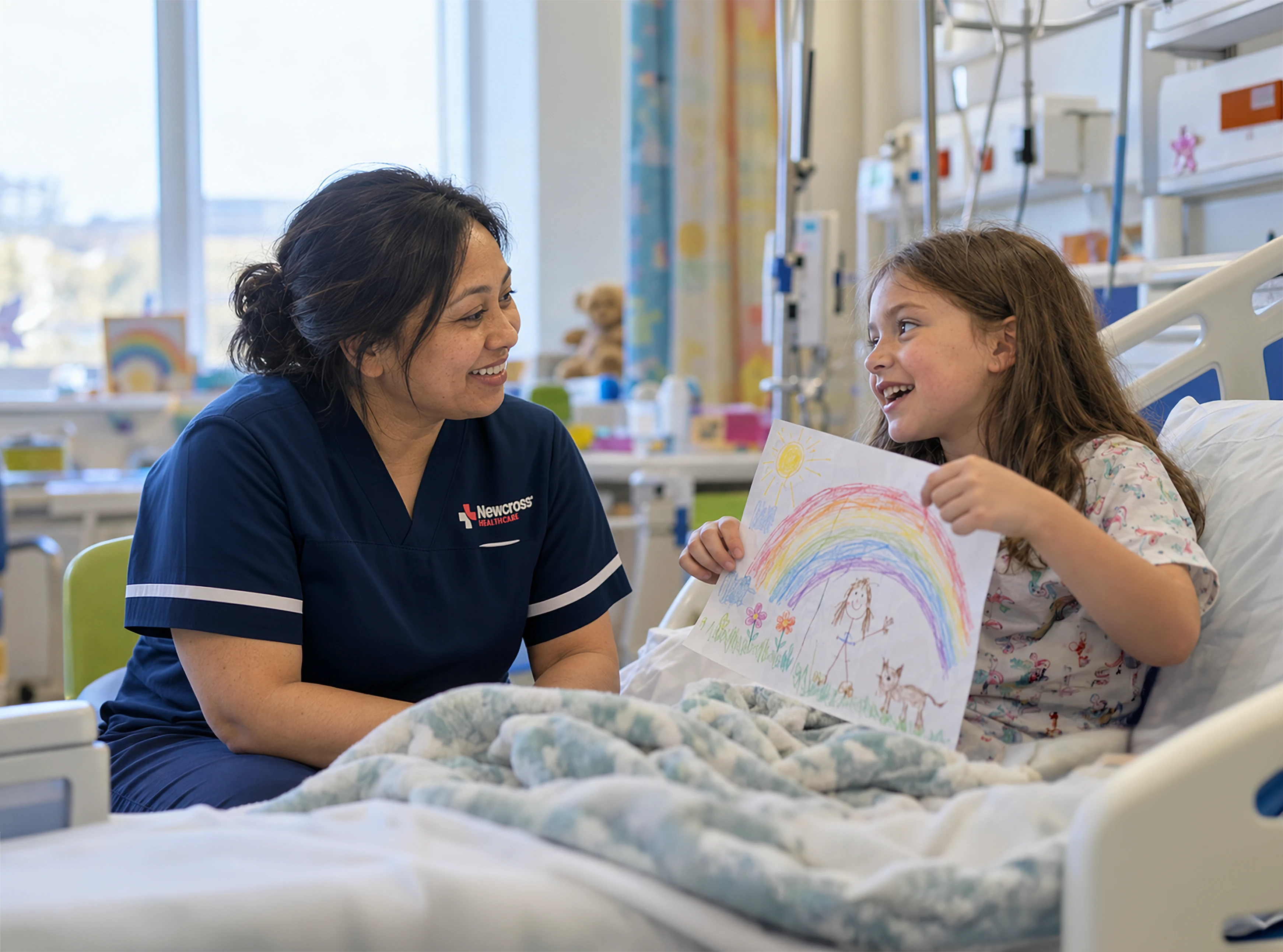 Asian British paediatric nurse in navy scrubs chatting with a young girl in a hospital bed as she shows the nurse her colourful drawing.
