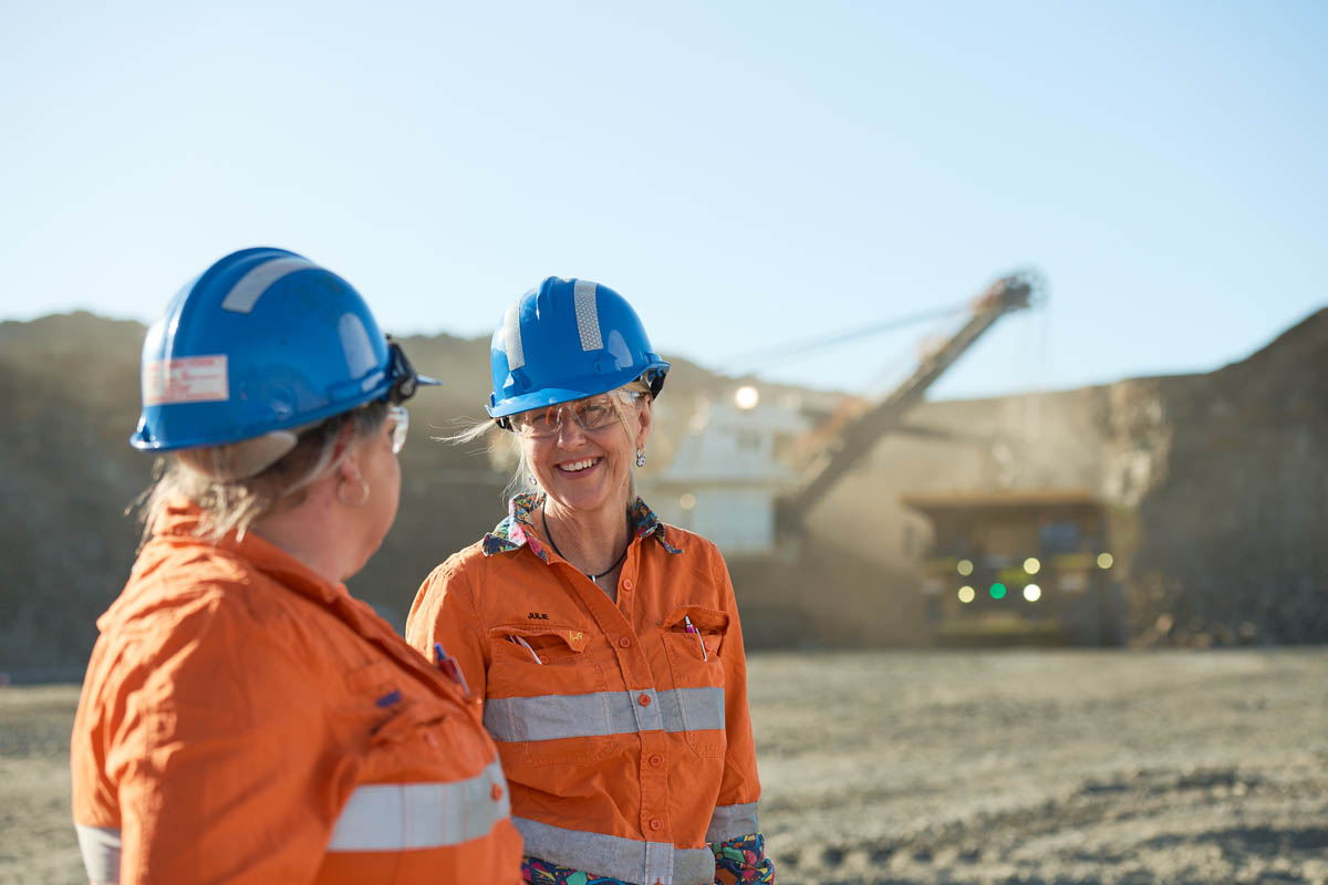 Two women in orange hi-vis PPE smiling at mine site with excavator background