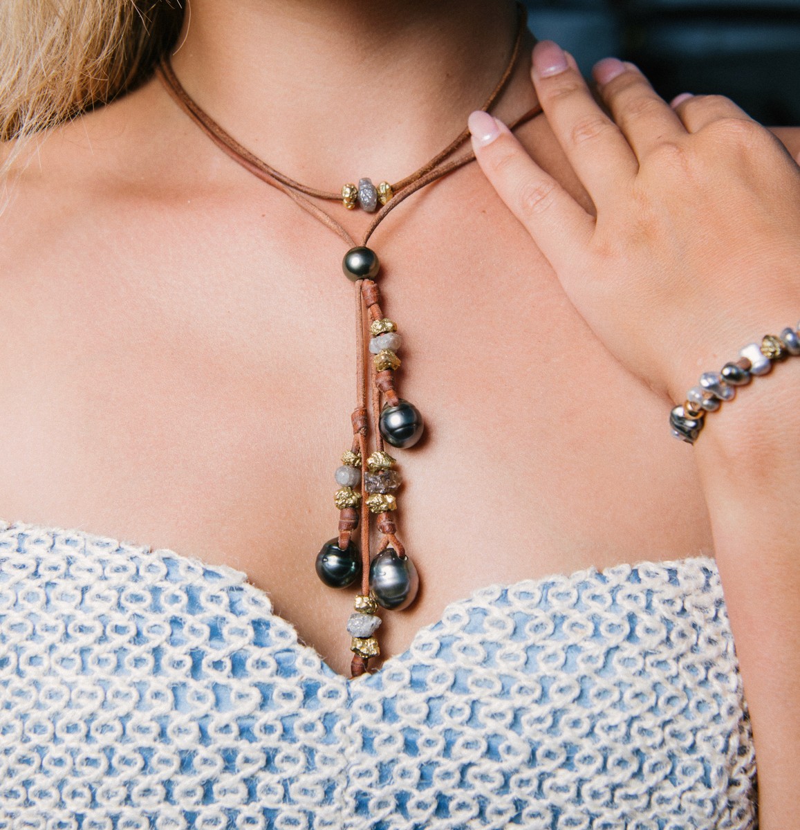 Woman wearing a luxurious Tahitian pearl necklace with raw gold accents and a matching bracelet, styled with an elegant blue and white woven top.