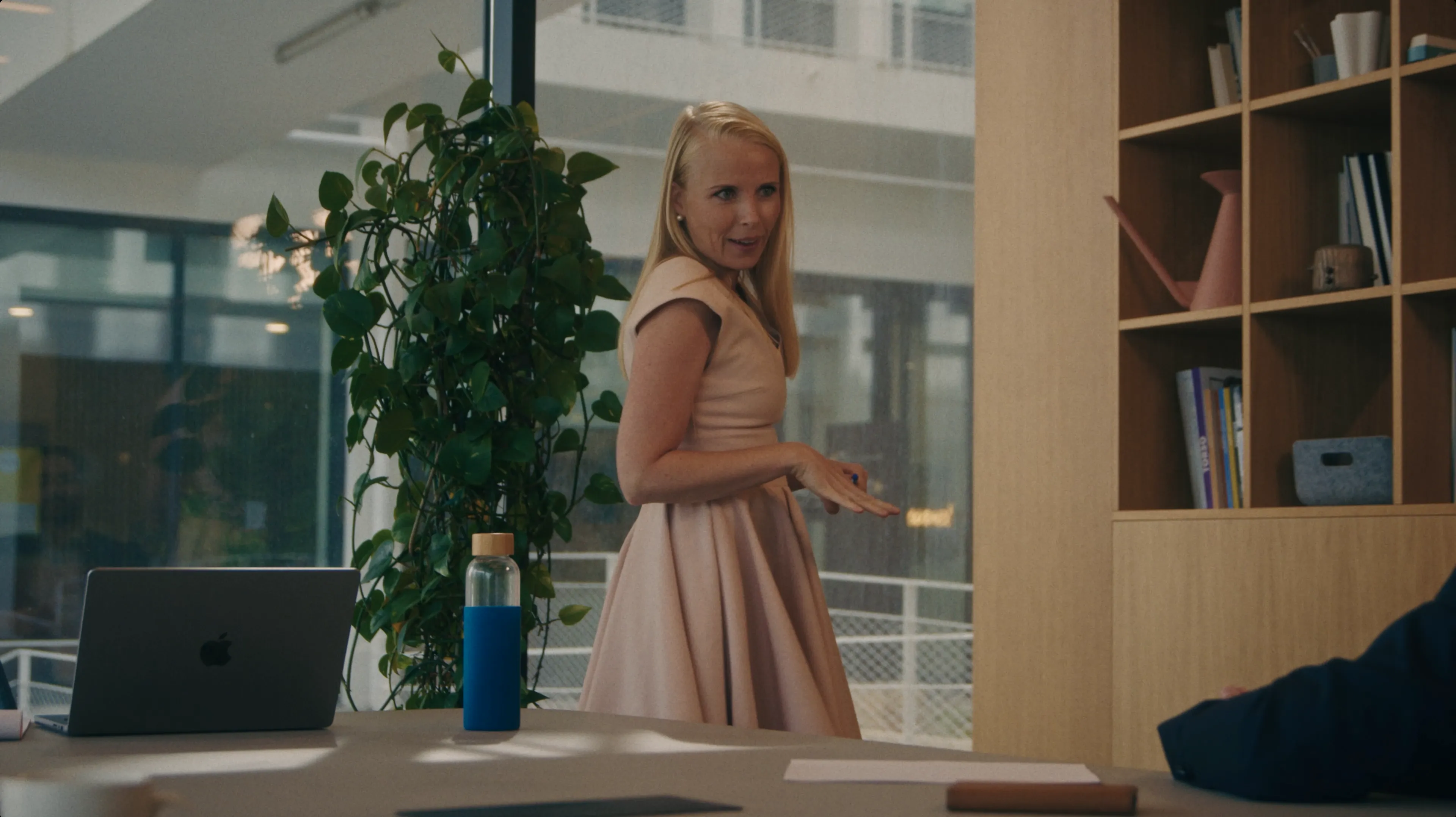 A woman in a light pink dress stands confidently in a modern office space with a leafy plant and bookshelves in the background, suggesting a professional yet creative environment.