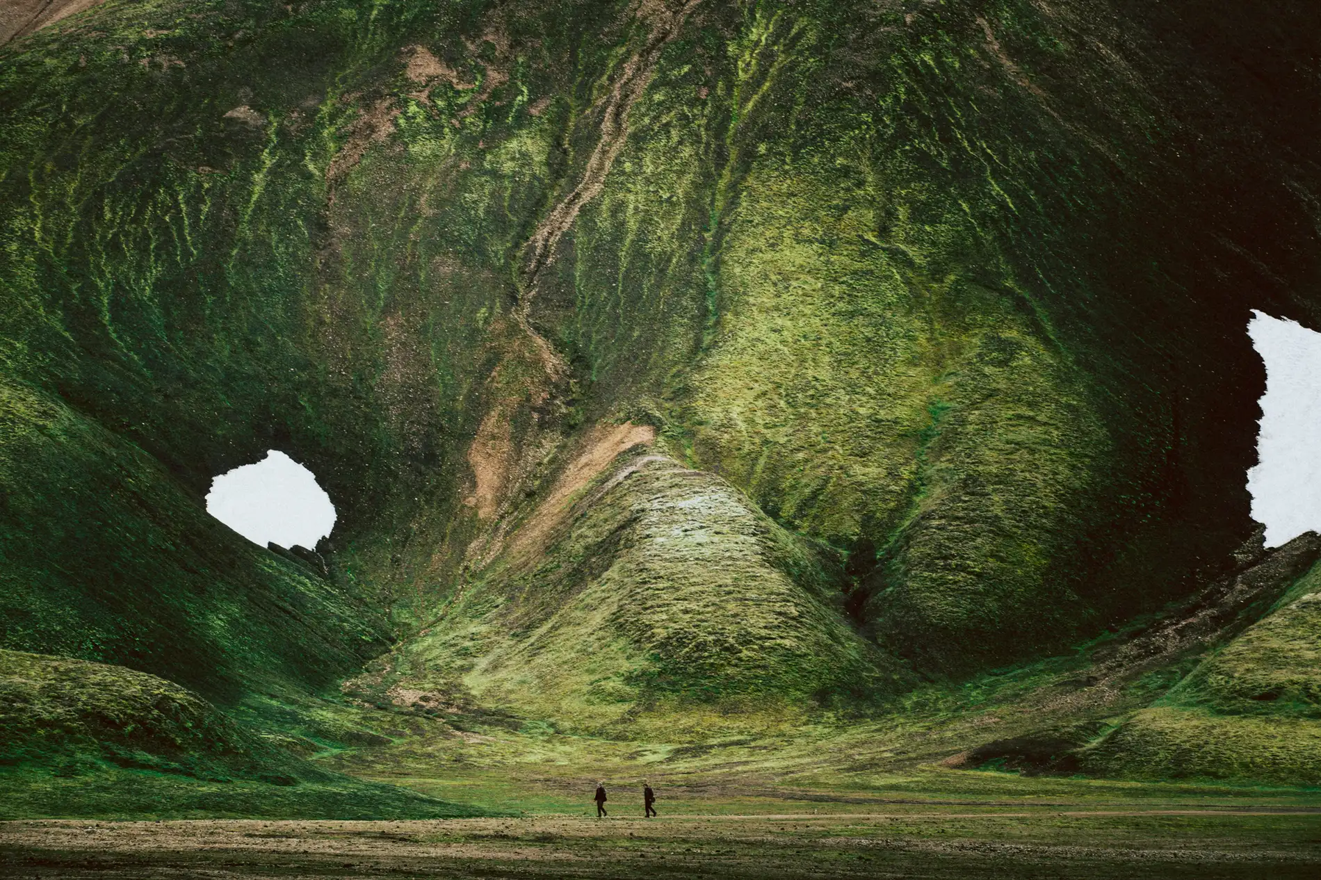 A green moss-covered volcanic crater with two small figures on the floor for scale