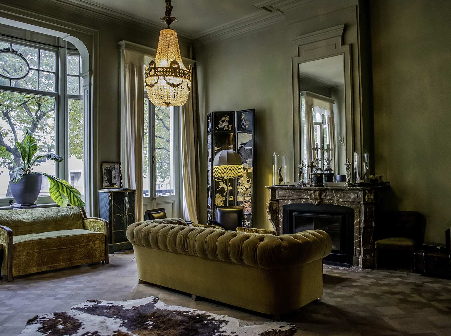 A lavish living room features gold velvet sofas, a crystal chandelier, and a marble fireplace under soft indoor lighting.