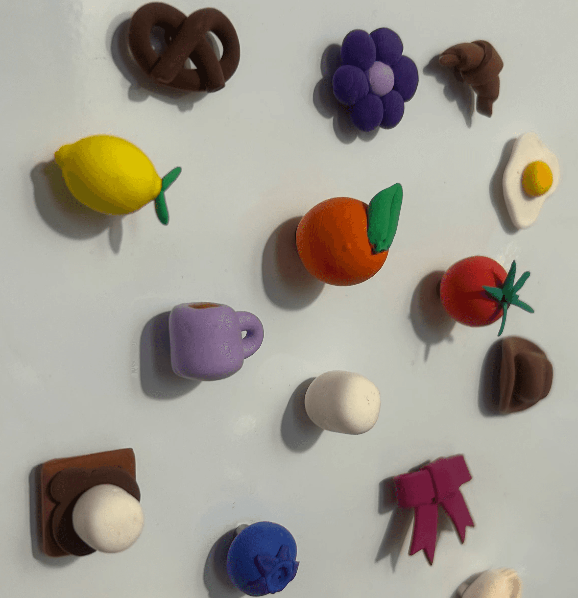 A collection of colorful, handmade clay magnets shaped like various items, including a pretzel, lemon, purple grapes, orange, coffee cup, tomato, and a sunny side up egg, displayed on a white magnetic surface.
