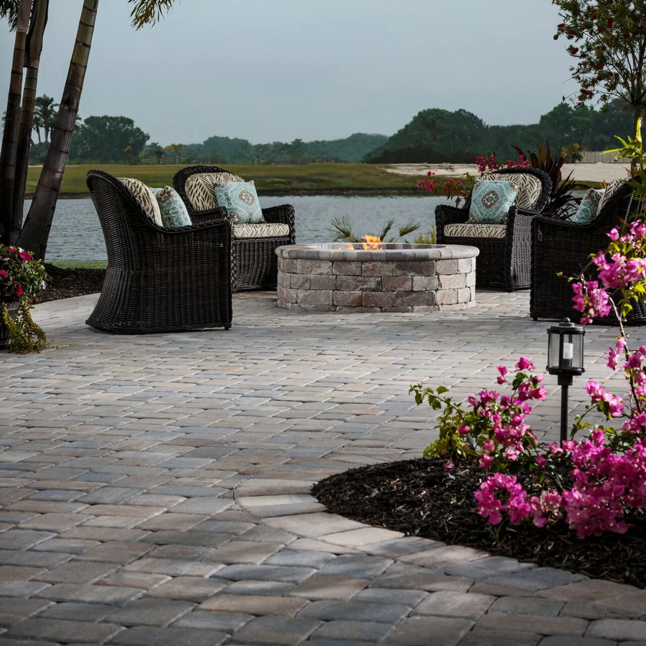 A relaxed outdoor setting Paver patio with woven chairs, a round fire pit and flowers beside a water view.