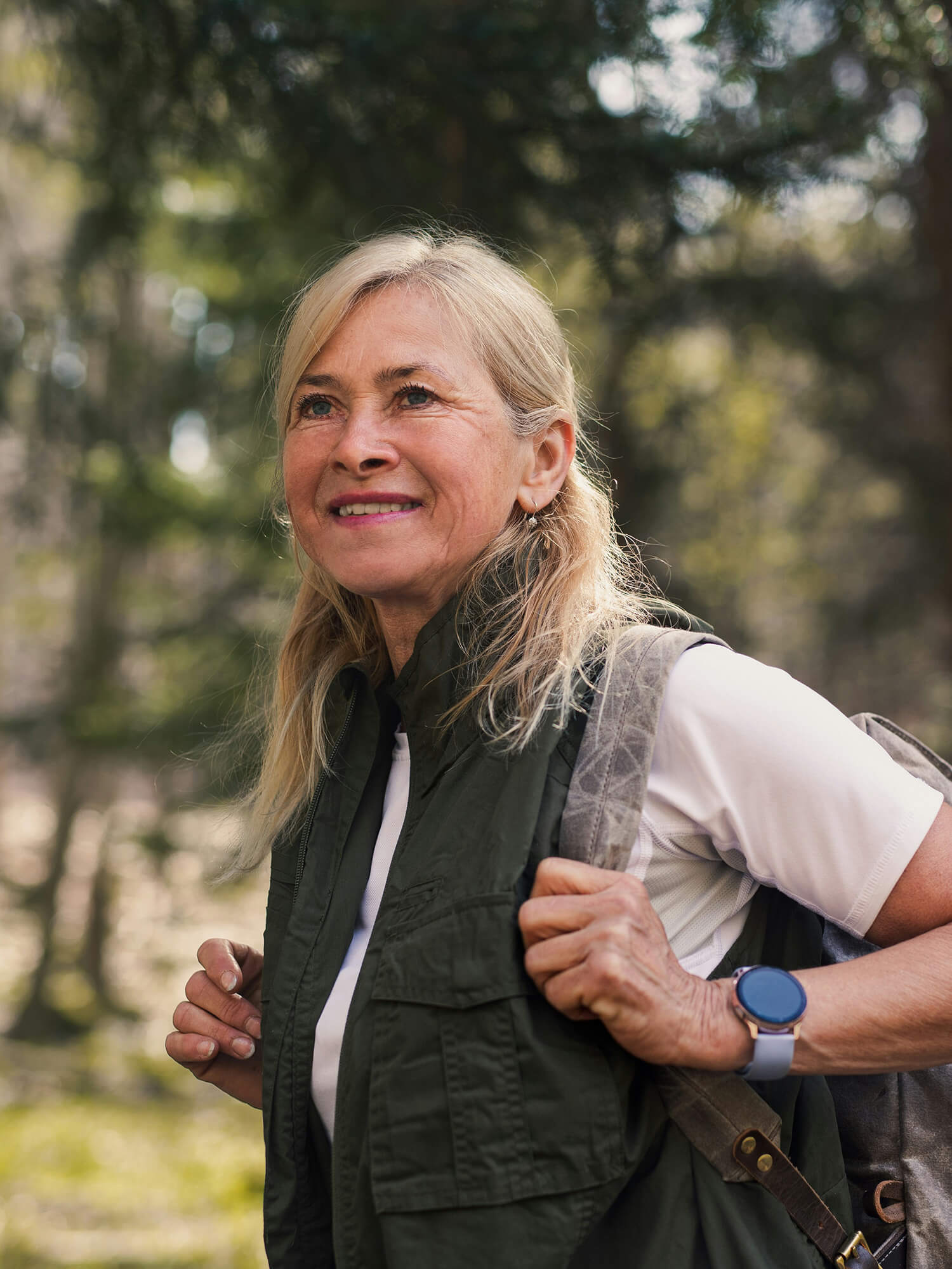 Middle-aged woman hiking in a forest, smiling and looking ahead, wearing a backpack, green vest, white shirt, and a smartwatch.