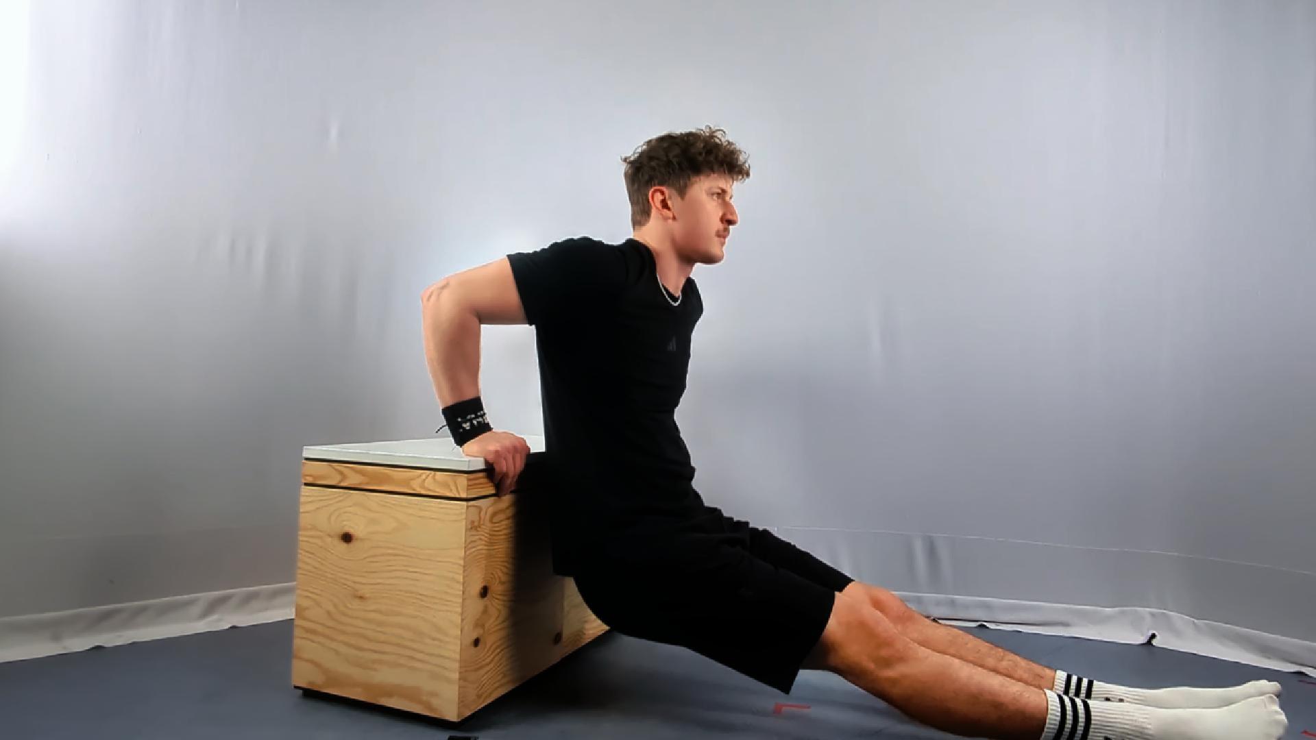 A man performs calisthenics bench dips in a home gym, using a wooden bench for support, wearing a black t-shirt and shorts, with his legs extended forward.