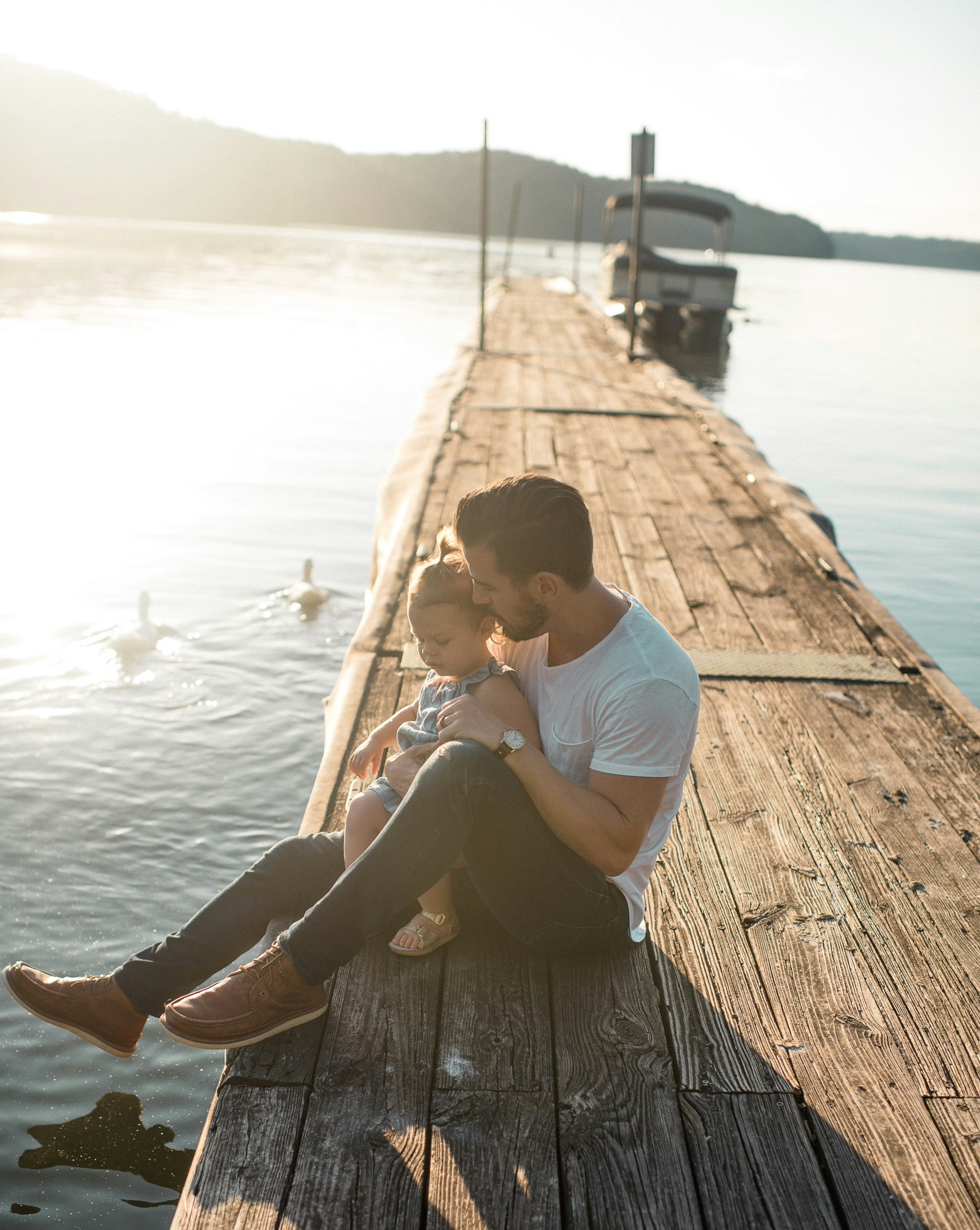 A man and a child sit together on a wooden dock by a serene lake with sunlight reflecting on the water and ducks swimming nearby.
