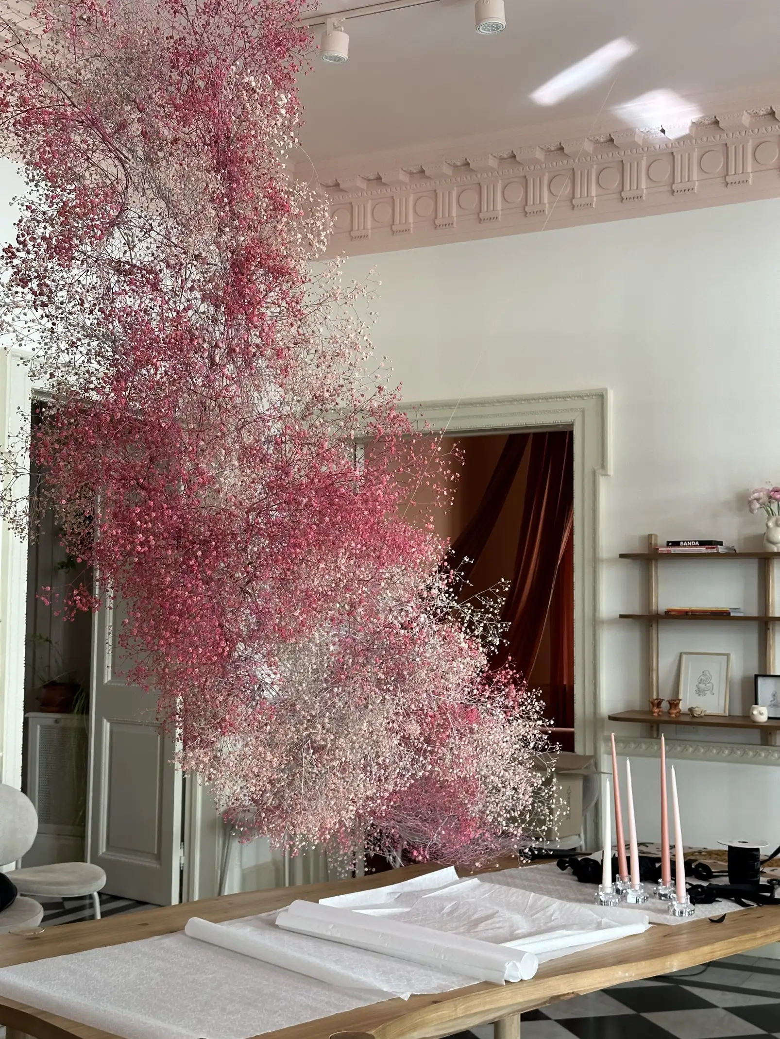 Arreglo suspendido de flores pequeñas en tonos rosados y blancos, creando un efecto de nube etérea sobre una mesa con velas rosas, con un fondo de sofás a cuadros y una pintura colorida en la pared.