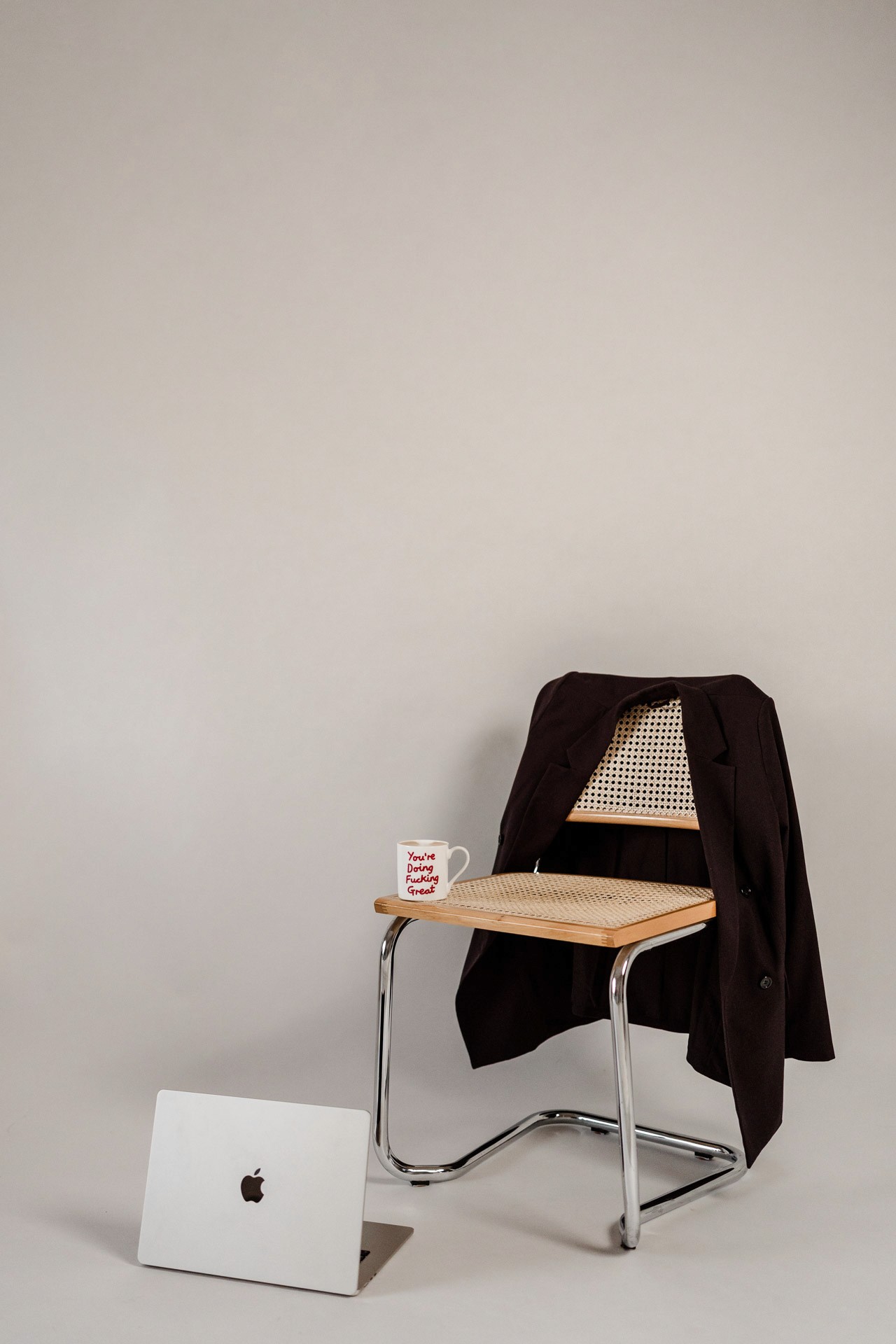 Korbstuhl mit Blazer über Rückenlehne und Tasse auf Sitzfläche steht vor hellem Hintergrund, auf dem Boden davor ein aufgeklappter Laptop 