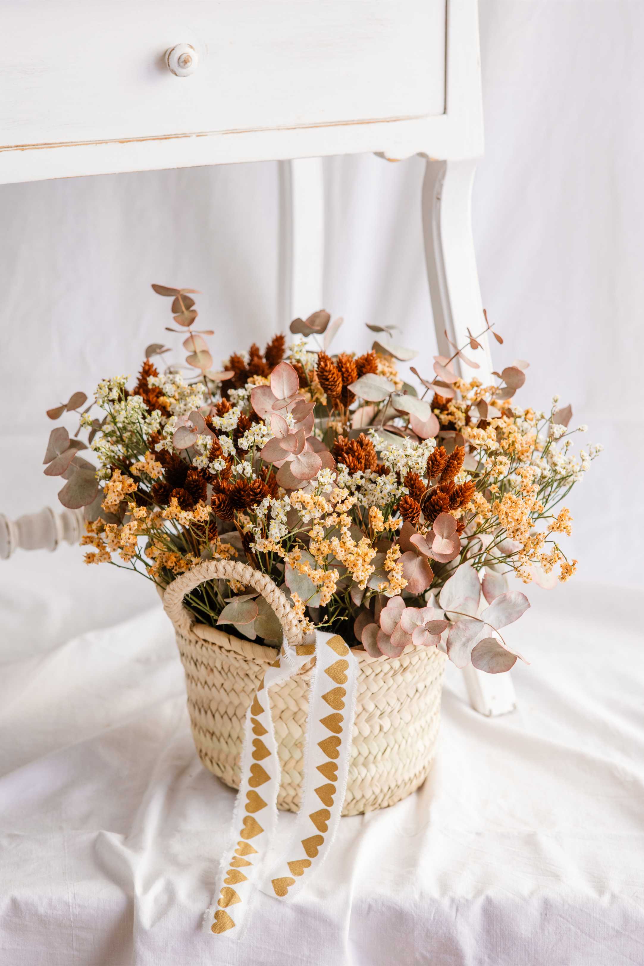 Cistell de vímet amb arranjament de flor seca en tons crema, rovellats i verds suaus, decorat amb cinta blanca amb cors daurats.