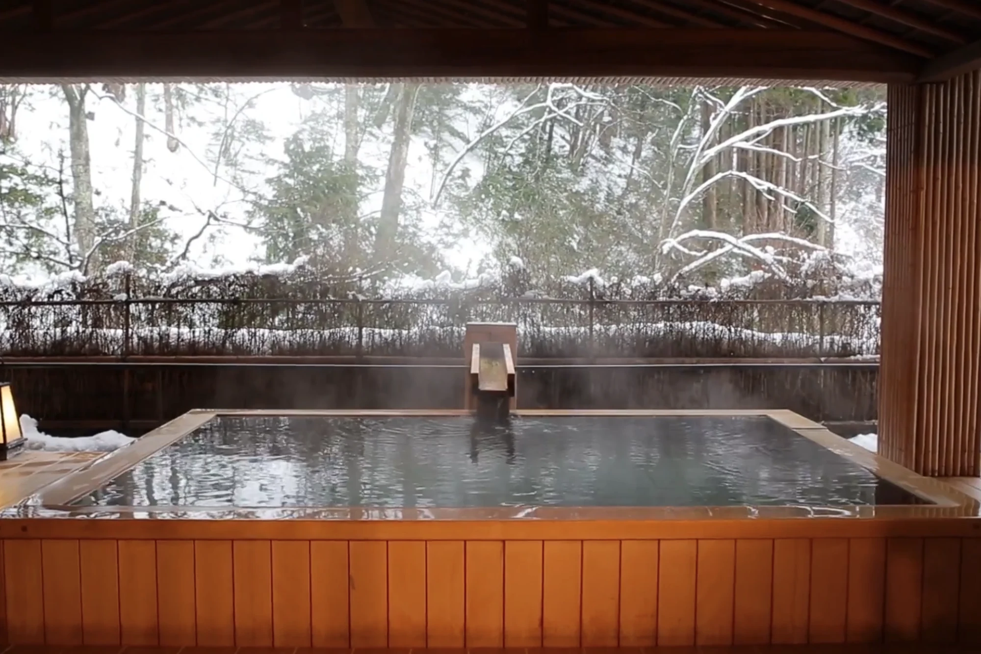 A timber-edged outdoor hot spring bath steaming in falling snow with snow-laden trees behind.