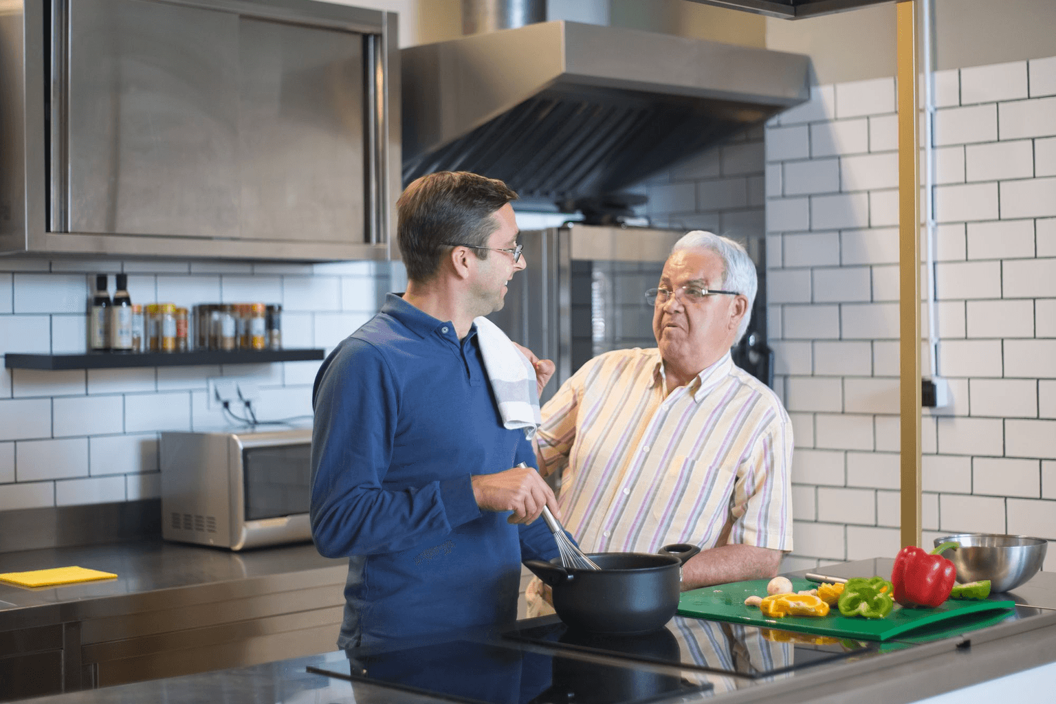 An older man cooking at the stove with his adult son, the two of them talking as they go.